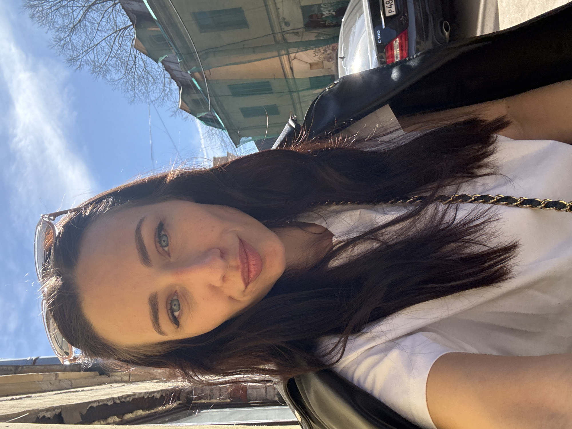 A woman with fair skin, blue eyes, and dark brown hair takes a selfie outdoors on a sunny day. She wears a white t-shirt and a black leather jacket. Sunglasses are perched on her head. A gold chain strap is visible across her chest. Buildings are visible in the background, along with a black car with license plate P48. The location is potentially Saint Petersburg, Russia.
