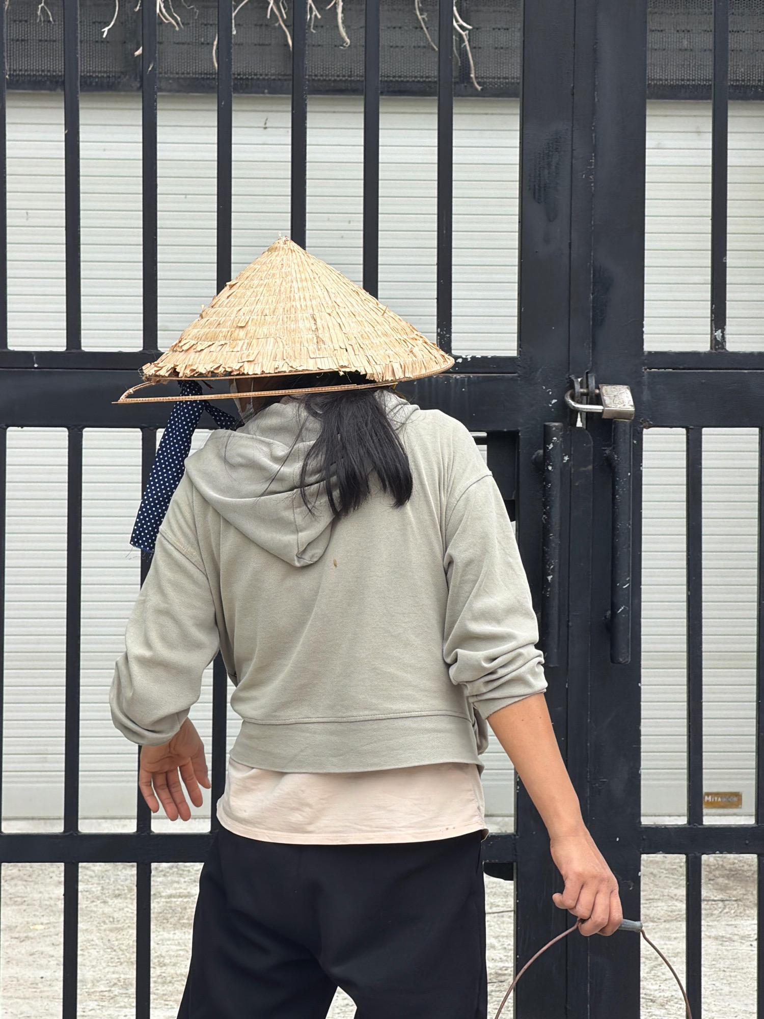 An individual wearing a conical straw hat and a light grey hoodie stands in front of a black metal gate with a padlock. The gate is partially closed, revealing a white corrugated metal surface behind it, likely a garage door or building facade. The individual's back is to the camera, and they are holding a wire handle, suggesting they are carrying a bucket or similar object. A piece of dark fabric with white polka dots is visible hanging from beneath the hat. The ground is paved with grey concrete. The scene appears to be outdoors in a built-up area of Nha Trang, Vietnam, possibly near a residence or commercial property.