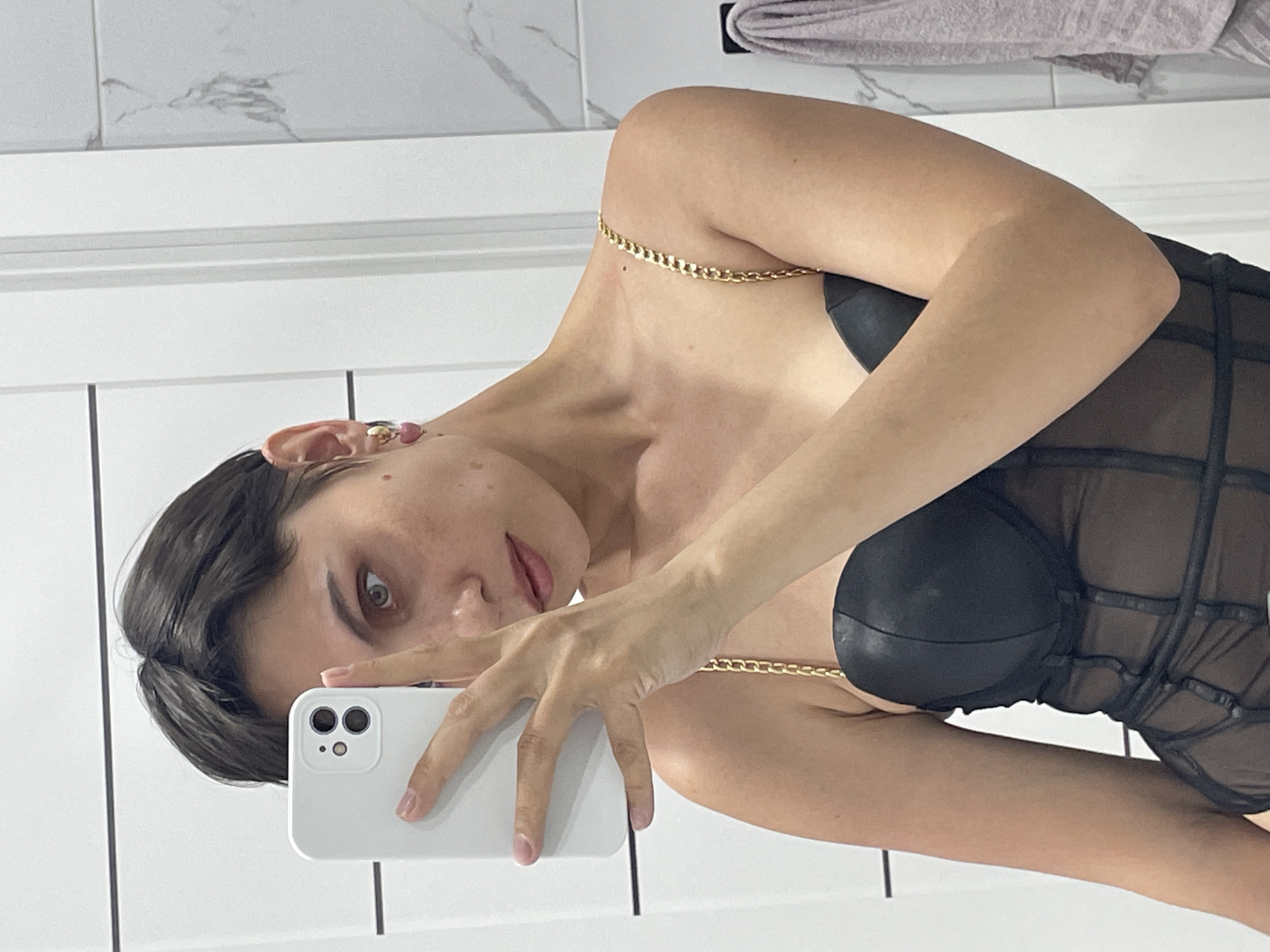 A woman is holding a white smartphone in her right hand, using it to take a selfie in what appears to be a bathroom. She is wearing a black corset-style top with gold chain straps. Her hair is short and dark. She is facing the camera with a slightly tilted head. The background consists of a white tiled wall and a towel hanging above. There is no indication in the image that it was taken in Izhevsk, Russia.