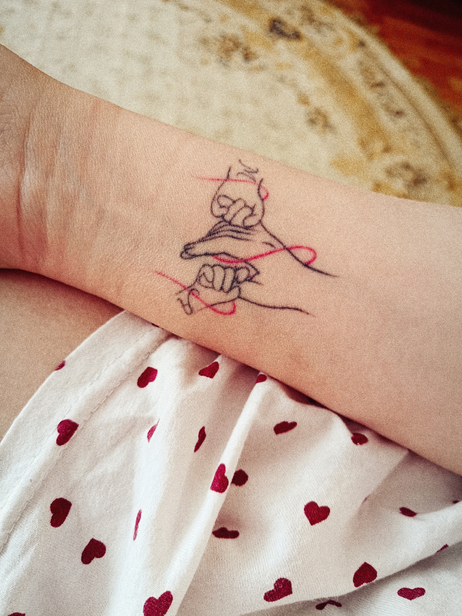 The image shows a forearm with a tattoo of two hands connected by a red string. The skin tone of the arm is light. The forearm is resting on a white fabric with red heart patterns. The background is blurry and appears to be a patterned rug. The location, Tobolsk, Russia, is not discernible from the image.