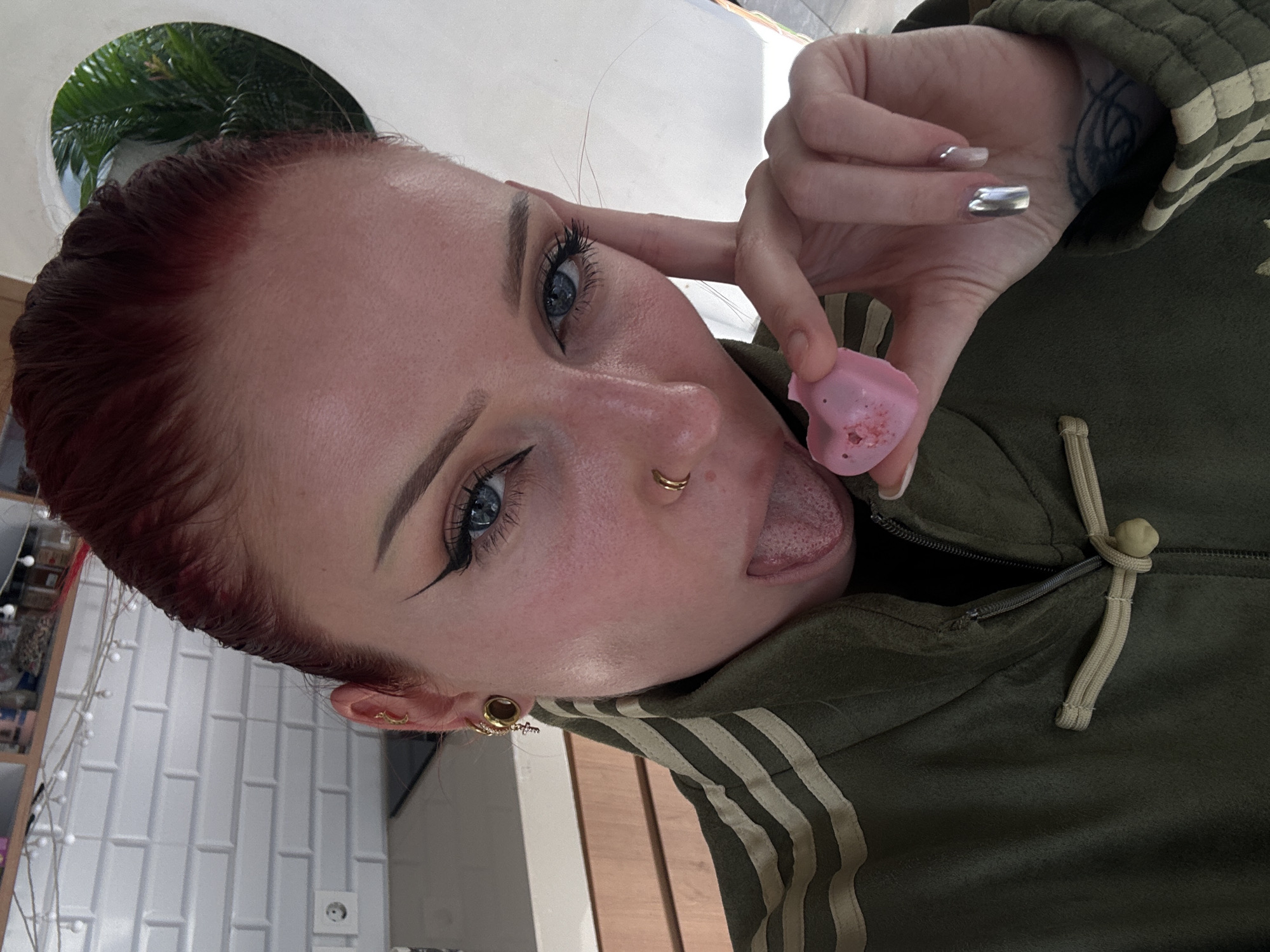 A fair-skinned person with red hair, prominent eyebrows, blue eyes with black eyeliner, and a gold nose ring is posing for a selfie. They are sticking out their tongue toward a pink, heart-shaped object held in their right hand. The person is wearing an olive-green hooded jacket with beige stripes. Tattoos are visible on their right wrist and hand. The background includes a white tiled wall, a window with a view of green foliage, and a partially visible shelf. The image appears to be taken indoors. While the person's location in North Kuta, Indonesia cannot be definitively determined from the image, the plant life visible through the window is consistent with a tropical location.