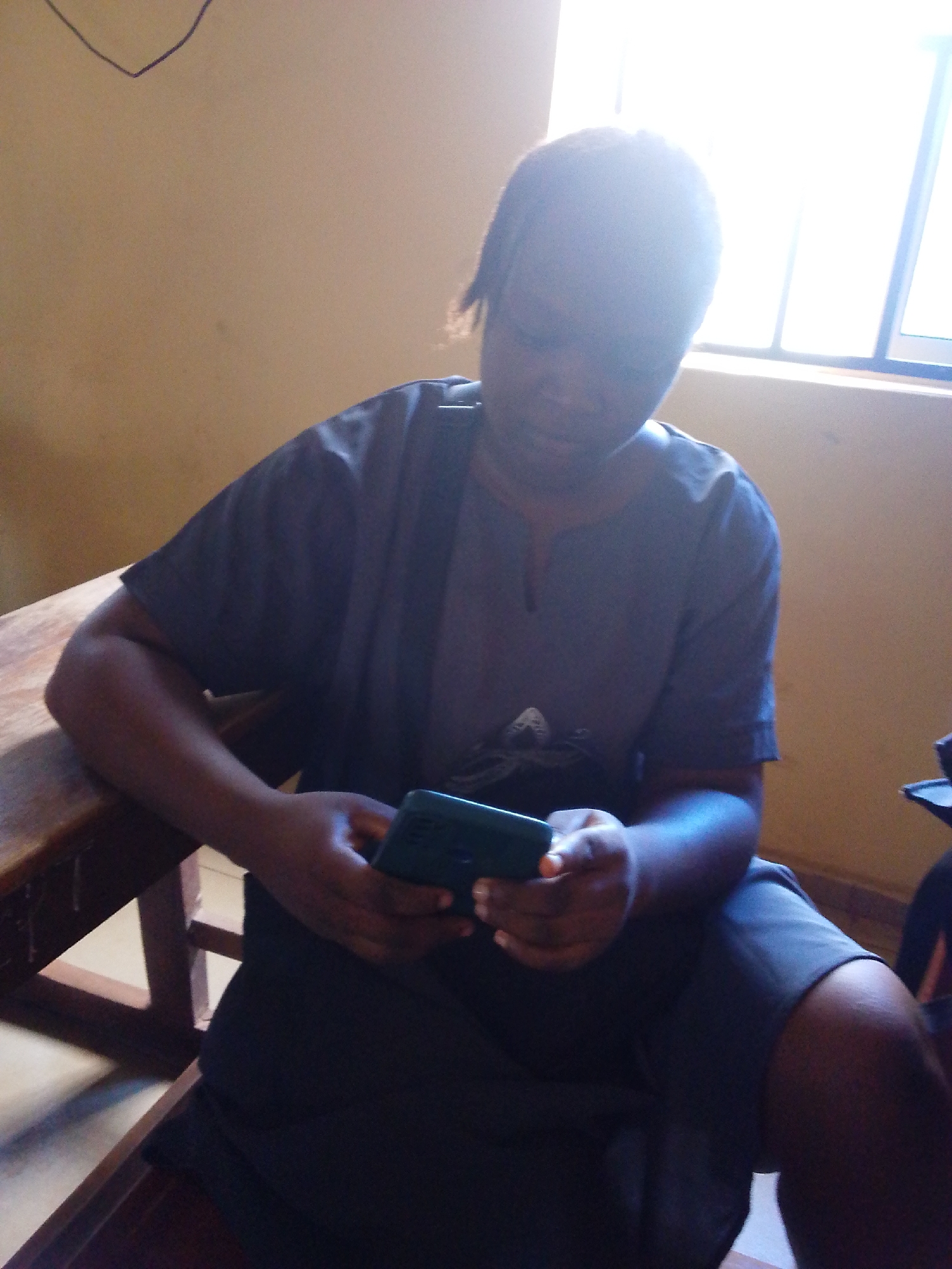 A person is seated on a wooden chair while looking at a mobile phone. The person is wearing a dark colored t-shirt and shorts. A window is visible behind the person. The location is Ukwu-Abwa, Nigeria.