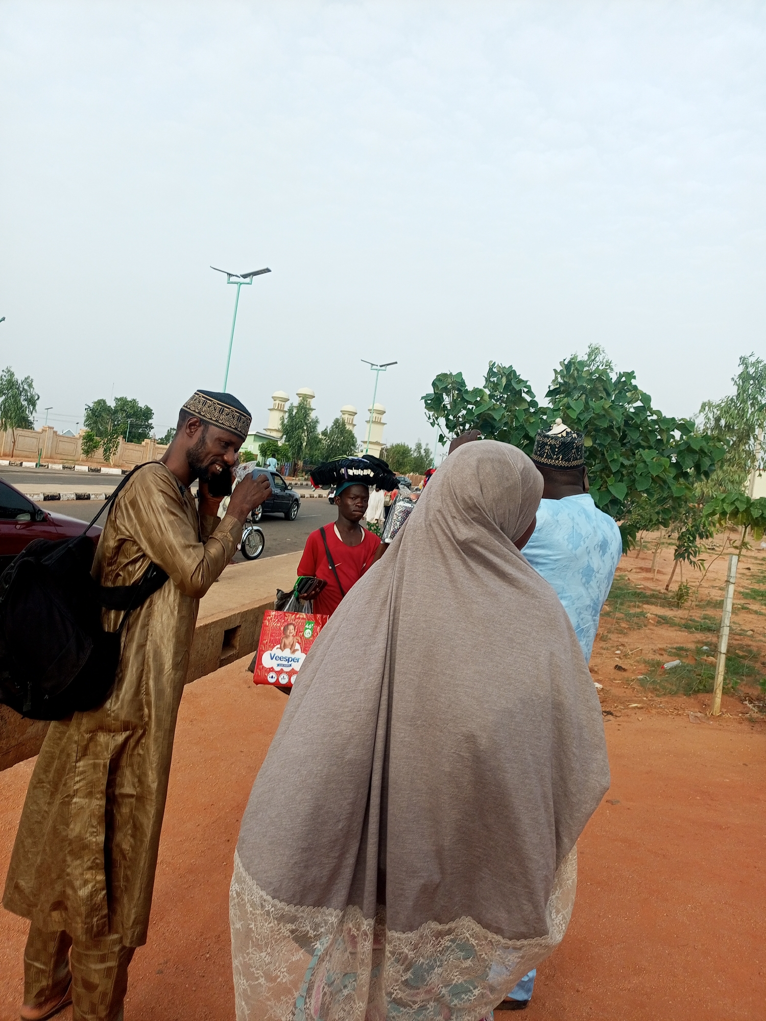 In Gombe, Nigeria, the image captures a street scene with several individuals. A man on the left, wearing a gold-colored traditional robe and cap, is using a cell phone. He has a black bag slung over his shoulder. To his right, another person, a woman, is wearing a tan-colored hijab that covers most of her body, obscuring her face. Beside her, another person is visible, wearing a light blue shirt and a cap similar in style to the first man's. Another man can be seen in a red shirt, with goods balanced on his head, and he carries a shopping bag with the word "Veesper" clearly printed on it.
In the background, there is a glimpse of a road with cars, a motorcycle, and mosque-like buildings. Streetlights are visible. The ground appears to be reddish. There are trees and shrubbery nearby. The sky is overcast.