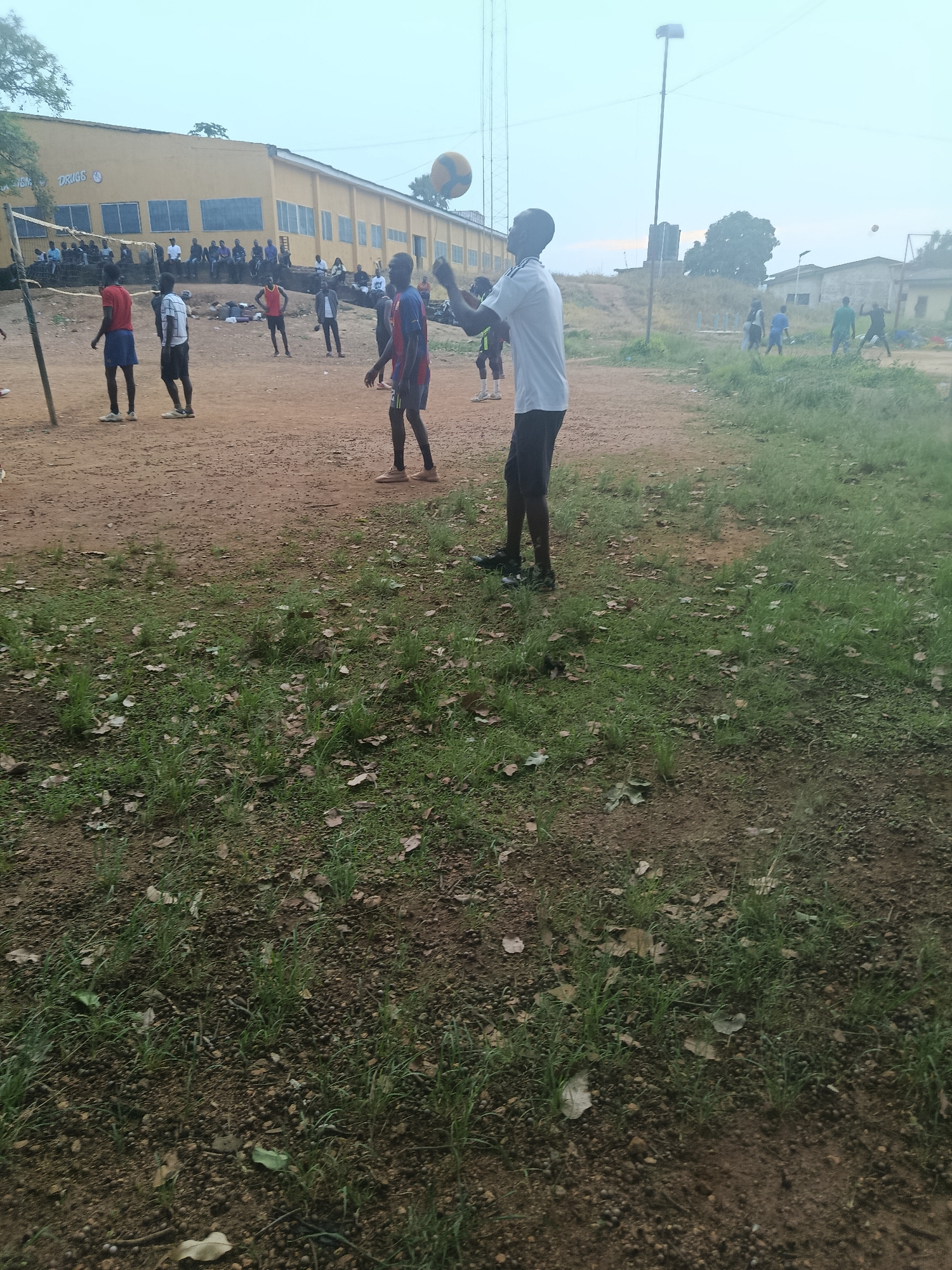 In Rafin Par, Nigeria, a group of young men are playing volleyball on a dusty court adjacent to a yellow building. The building, which appears to be a school or community center, has the words "DRUGS" and "DRUG" visible on its facade. In the foreground, a young man in a white t-shirt and black shorts is poised to hit the ball, while other players in various athletic attire are scattered across the court. The environment is characterized by dry, reddish-brown earth, patches of sparse green grass, and fallen leaves. In the background, several onlookers are seated on a low wall in front of the building, observing the game. The sky is overcast, suggesting a cloudy day.