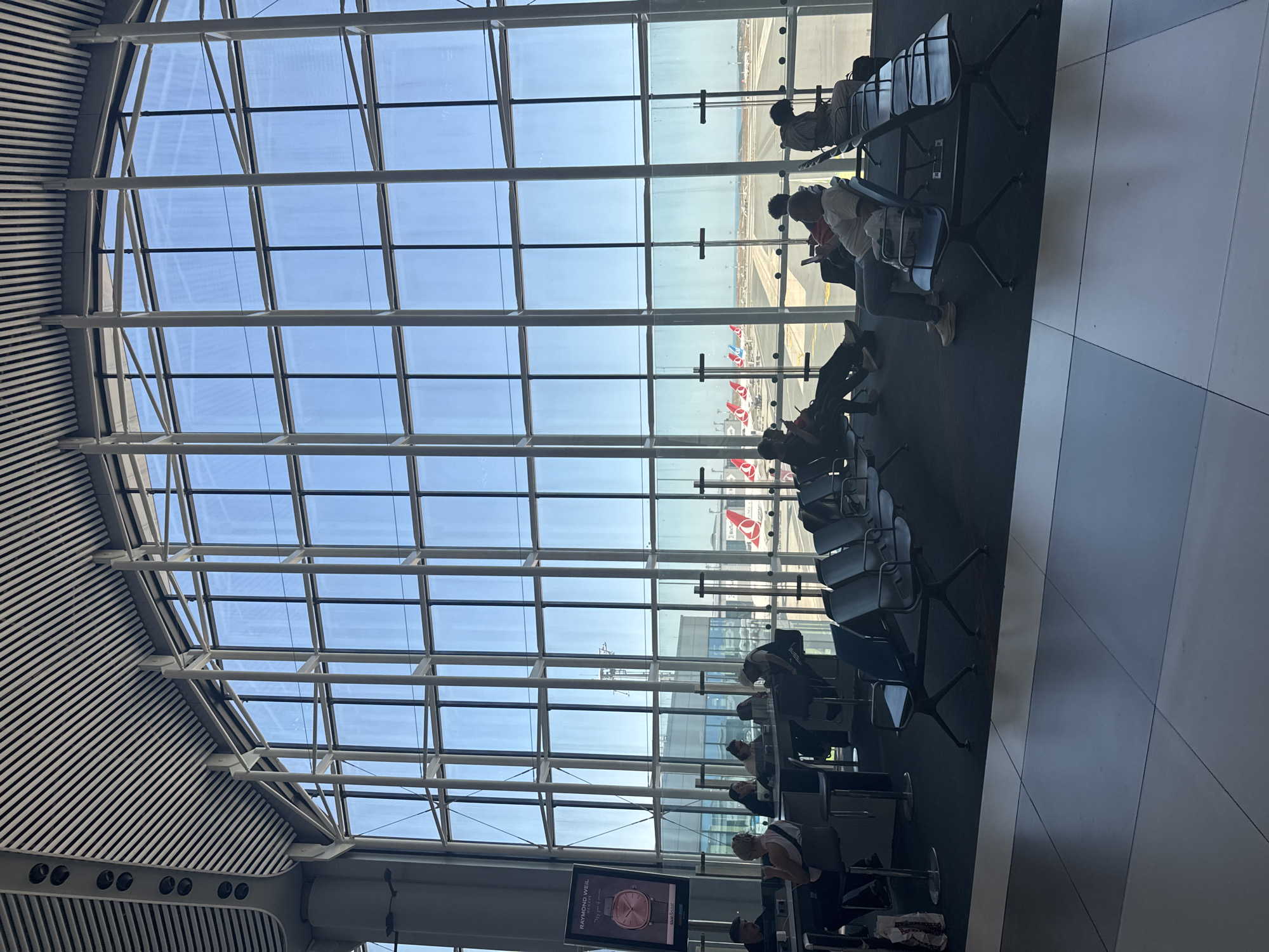 The image depicts a waiting area at Istanbul Airport in Arnavutköy, Turkey. A large, floor-to-ceiling window offers a view of an airport tarmac, where several airplanes with Turkish Airlines livery are visible. Inside the waiting area, rows of dark, modern airport seating are arranged. Several individuals are seated, some engaged with their phones, others looking out the window or resting. In the foreground, the tiled floor of the airport terminal is visible. To the left, a digital advertisement for Raymond Weil watches is displayed on a tall screen. The architectural style of the terminal features a high, vaulted ceiling with a grid-like structure. The overall scene suggests a typical airport environment, characterized by waiting passengers and the expansive glass facade overlooking the active tarmac.