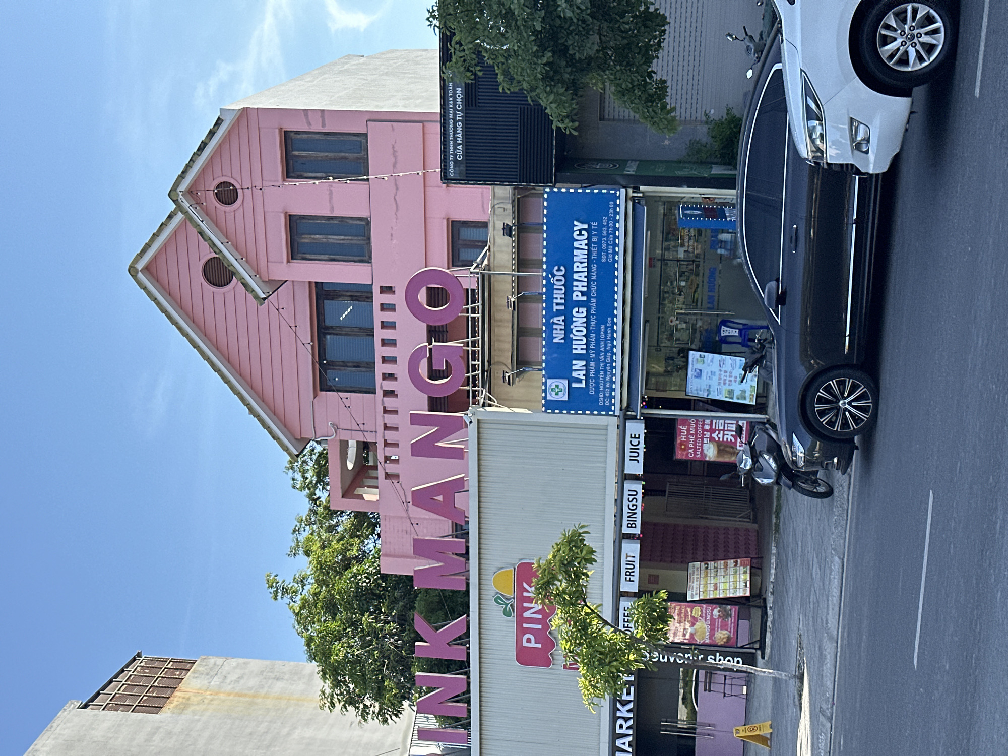 The image depicts a street scene in Đà Nẵng, Vietnam, featuring a pink building with the signage "MANGO" on the upper part and "PINK MARKET" below. The establishment offers "FRUIT," "BINGSU," and "JUICE." Next to it is "LAN HƯƠNG PHARMACY," displaying information in Vietnamese, including opening hours and contact details. A partially visible black and white car occupies the right side of the frame, along with a motorbike. The street pavement and road markings are visible. Signage for "Souvenir shop," "COFFEE" and "salted Coffee" are also on the front of the establishment.