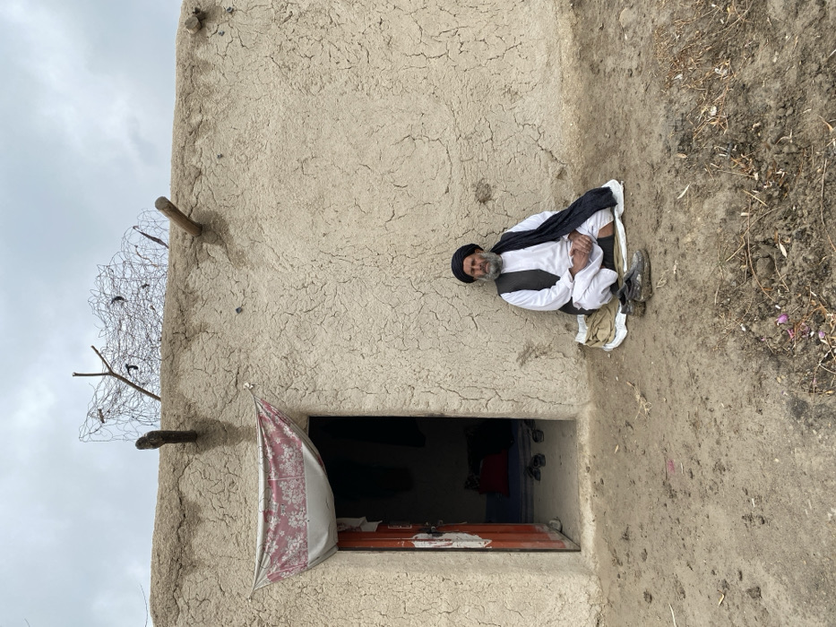 An elderly man with a black turban and white clothing sits outside a mud-brick building. He's positioned to the right of a small, low doorway. A patterned cloth hangs partially over the doorway. A makeshift fence with barbed wire is visible on the left side of the building. The ground around the building is bare, with some sparse vegetation to the right of the man. The sky is overcast.