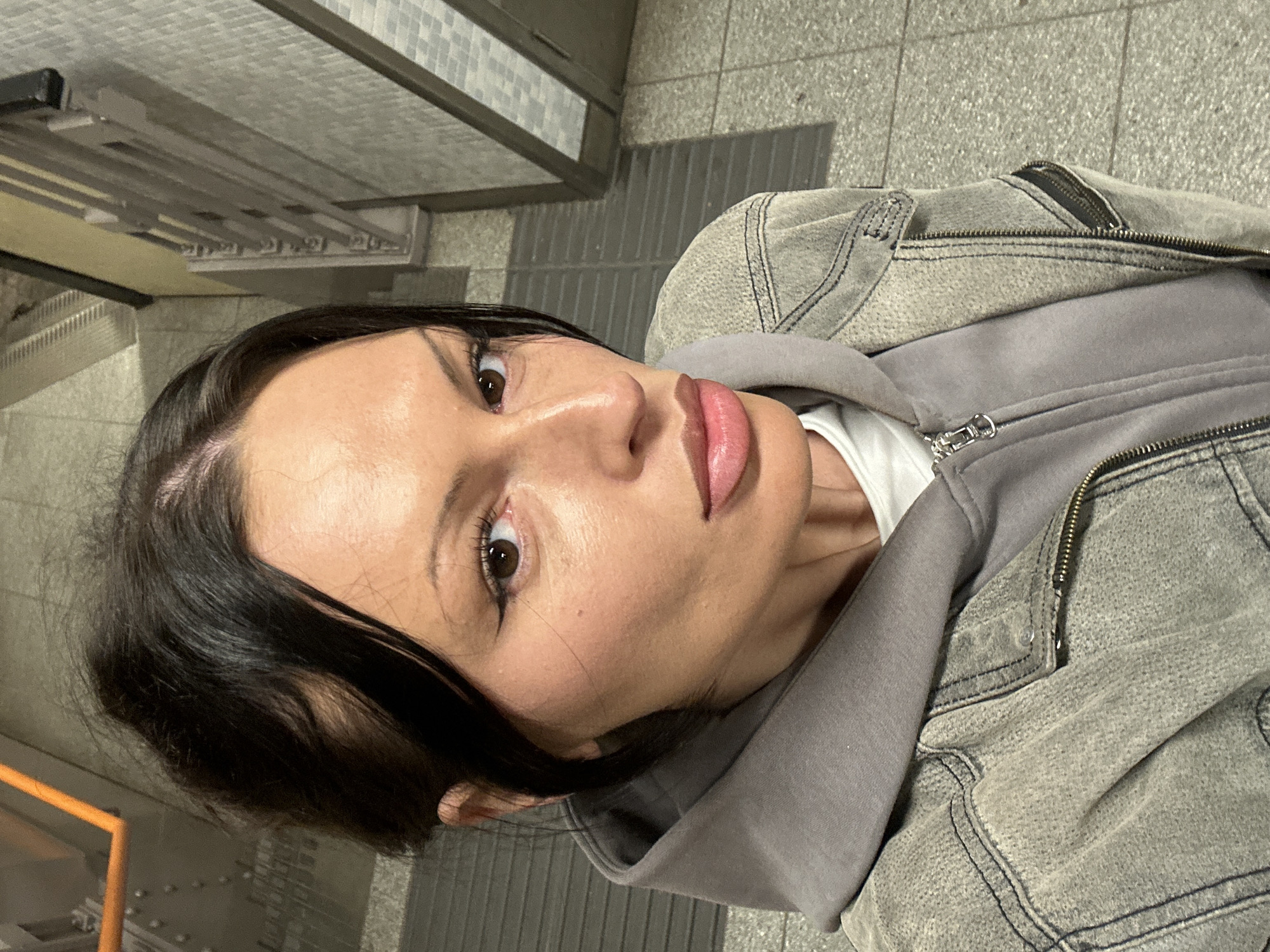 A person with dark, parted hair and visible eye makeup, including winged eyeliner, along with lip gloss, looks directly at the camera, capturing a self-portrait. They are wearing a light grey denim-style jacket over a grey zippered hoodie, which covers a white t-shirt. A small mole is present on the subject's right cheek. The background depicts an interior public space consistent with a London Underground station in the city of London, United Kingdom. Environmental elements include light grey tiled flooring, some sections featuring darker grey textured patterns, and light-colored square-tiled walls. Structural components of a stairway or escalator, including dark railings and a section of an orange handrail, are visible in the upper background.