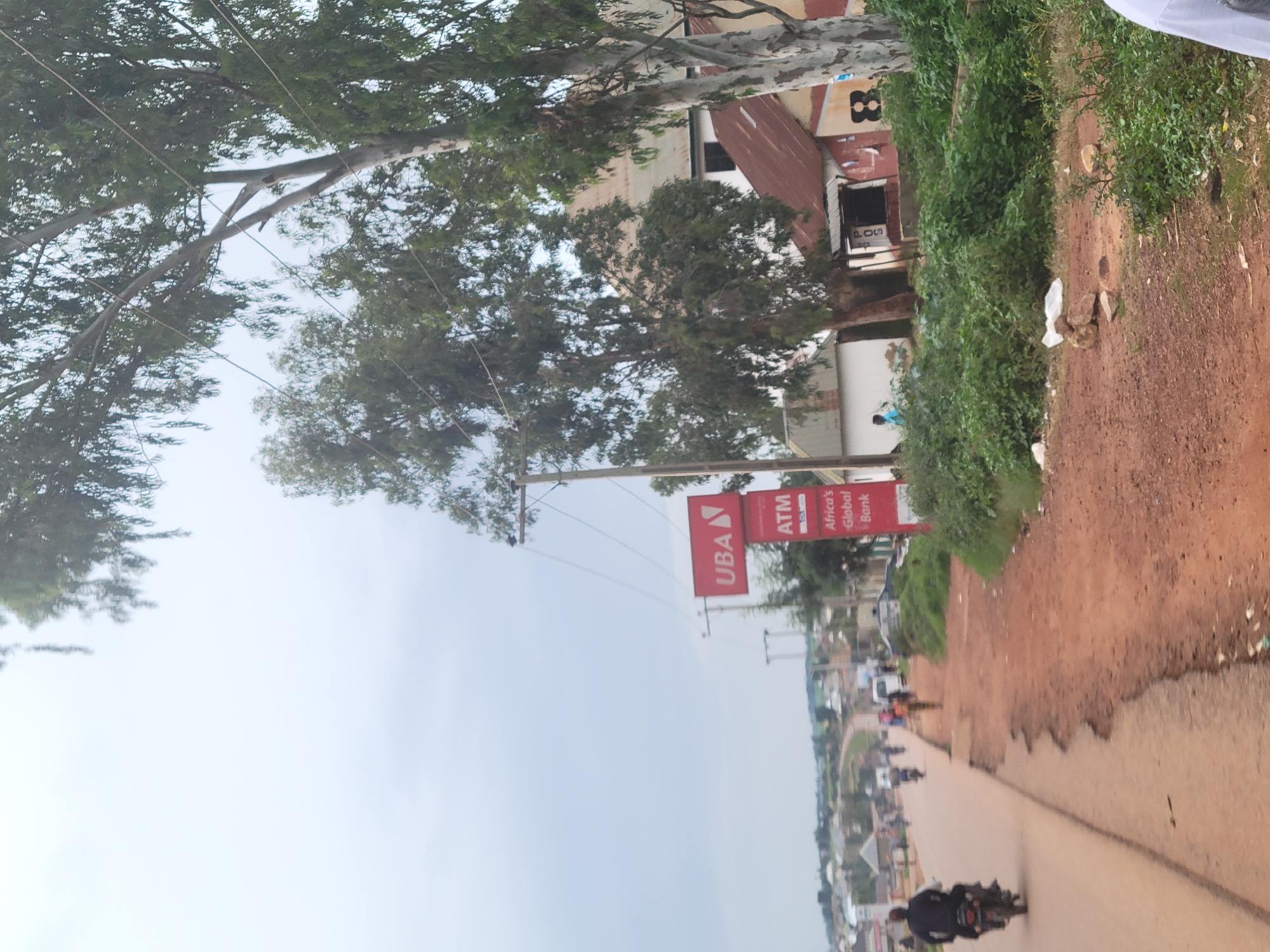 The image depicts a roadside scene in Bokkos, Nigeria, likely on a cloudy day. A paved road, with some visible cracks and disrepair, curves to the left and recedes into the distance. Several motorcycles are present on the road; one in the foreground is ridden by a person wearing a dark shirt, while others are further down the road. A person in a light blue shirt is visible standing on the side of the road.
To the right of the road, there is a red sign with "UBA" and "ATM" written on it, indicating a bank. Adjacent to the sign are several buildings with red and white corrugated iron roofs, interspersed with greenery. Large trees with sparse foliage line the area between the road and the buildings. Some debris, including rocks and white plastic bags, are scattered on the unpaved area to the right of the road. The background shows a slightly hazy view of more buildings and hilly terrain.