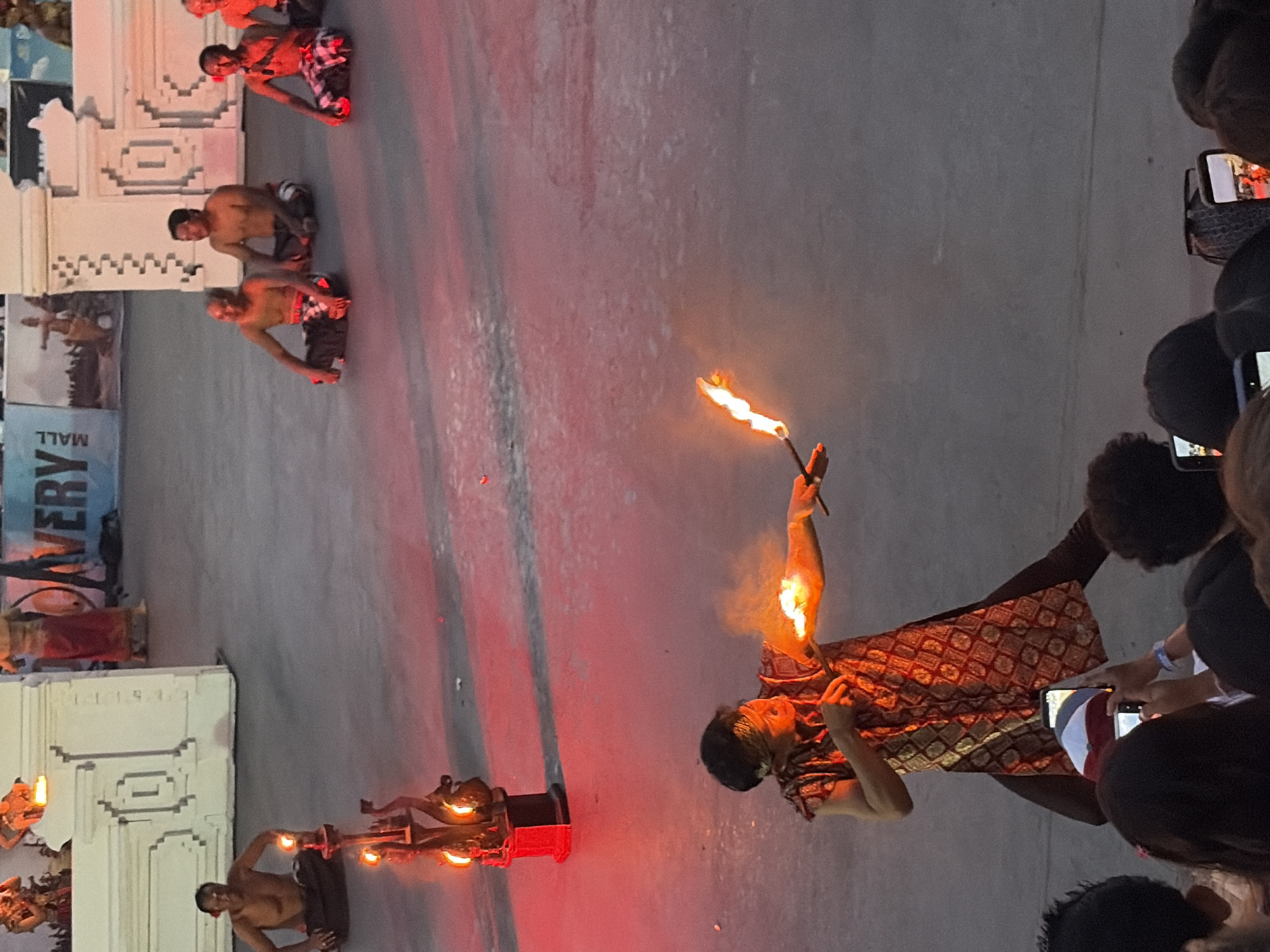 This image captures a vibrant cultural performance in Kuta, Indonesia. The scene is set outdoors at dusk, with the warm glow of fire illuminating the performers and the surrounding area. The primary subjects are a group of men participating in a fire-dancing or fire-eating ritual. They are dressed in traditional Indonesian attire, including patterned sarongs.
One performer in the foreground is actively engaged in the performance, holding a flaming torch and exhibiting a focused expression. His movements suggest a dynamic and potentially dangerous act. Other performers are visible in the background, also interacting with fire, either holding torches or positioned near fire elements.
The setting includes what appears to be a decorative stage or backdrop with intricate architectural details, possibly representing a temple or traditional structure. In the upper left, a banner with the word "MALL" is visible, suggesting the performance is taking place near a commercial area. Spectators are gathered on the right side of the image, many holding up their mobile phones to record the event, indicating a popular public spectacle. The lighting suggests it is either early evening or late afternoon, with artificial lights and the fire contributing to the overall illumination. The atmosphere seems to be one of excitement and cultural immersion.
