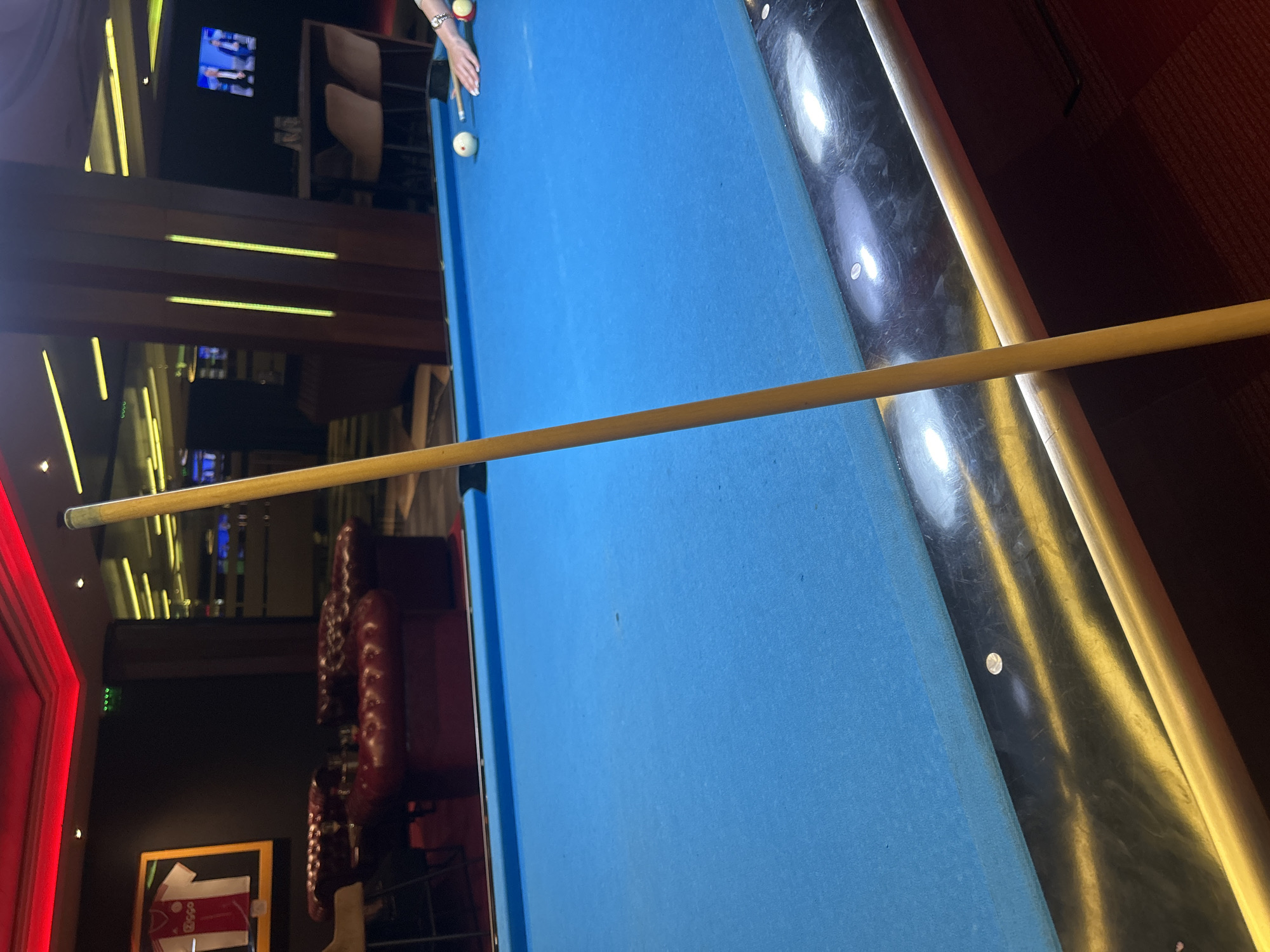 The image depicts a billiard table in a dimly lit environment. A person's hand, wearing a watch, is visible near two billiard balls on the light blue table surface. A cue stick lies across the table, pointing towards the left side of the frame. The table has a dark-colored rail with shiny, circular inlays.
In the background, there's a red leather sofa and a framed jersey on the wall. The setting appears to be a bar or entertainment area. The location is likely Sofia, Bulgaria, based on the frame of the jersey on the wall in the background.