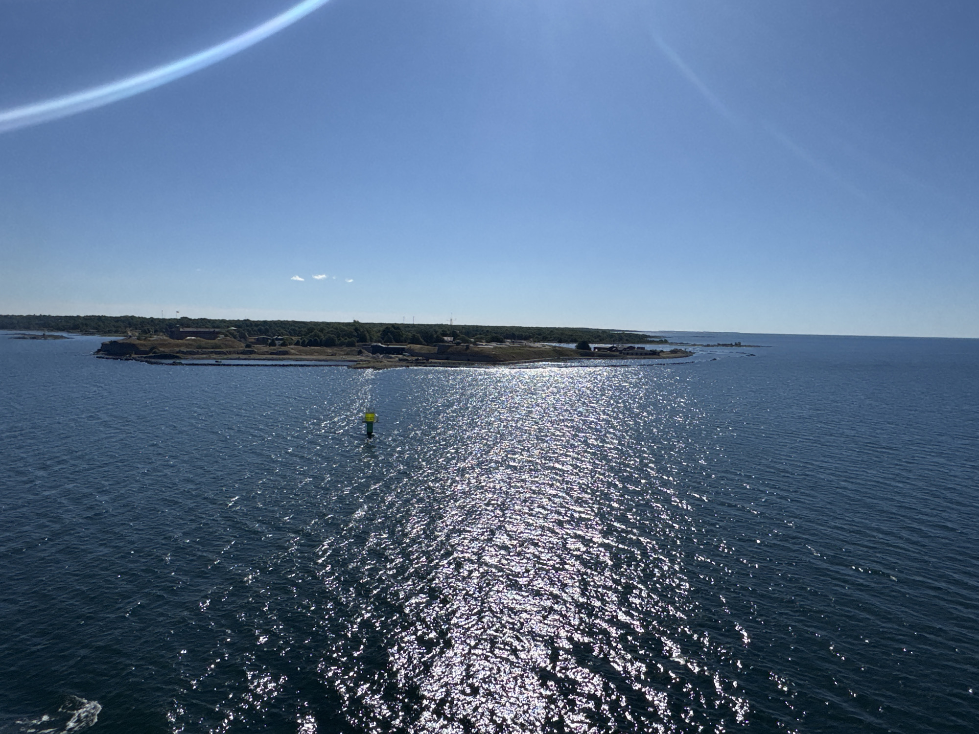 The image shows a view of the sea towards the island of Drottningskär, Sweden. The sky is clear blue with a bright sun creating a lens flare effect in the upper left corner. A long, thin cloud or atmospheric phenomenon stretches across the sky. The sea is dark blue with the sun's reflection creating a shimmering, sparkly surface. In the distance, the island of Drottningskär is visible, characterized by its rocky terrain and a cluster of buildings, possibly part of a fortress or historical site. A green and yellow buoy is present in the water. The overall scene depicts a calm, sunny day at sea with a view of the Swedish coastline.