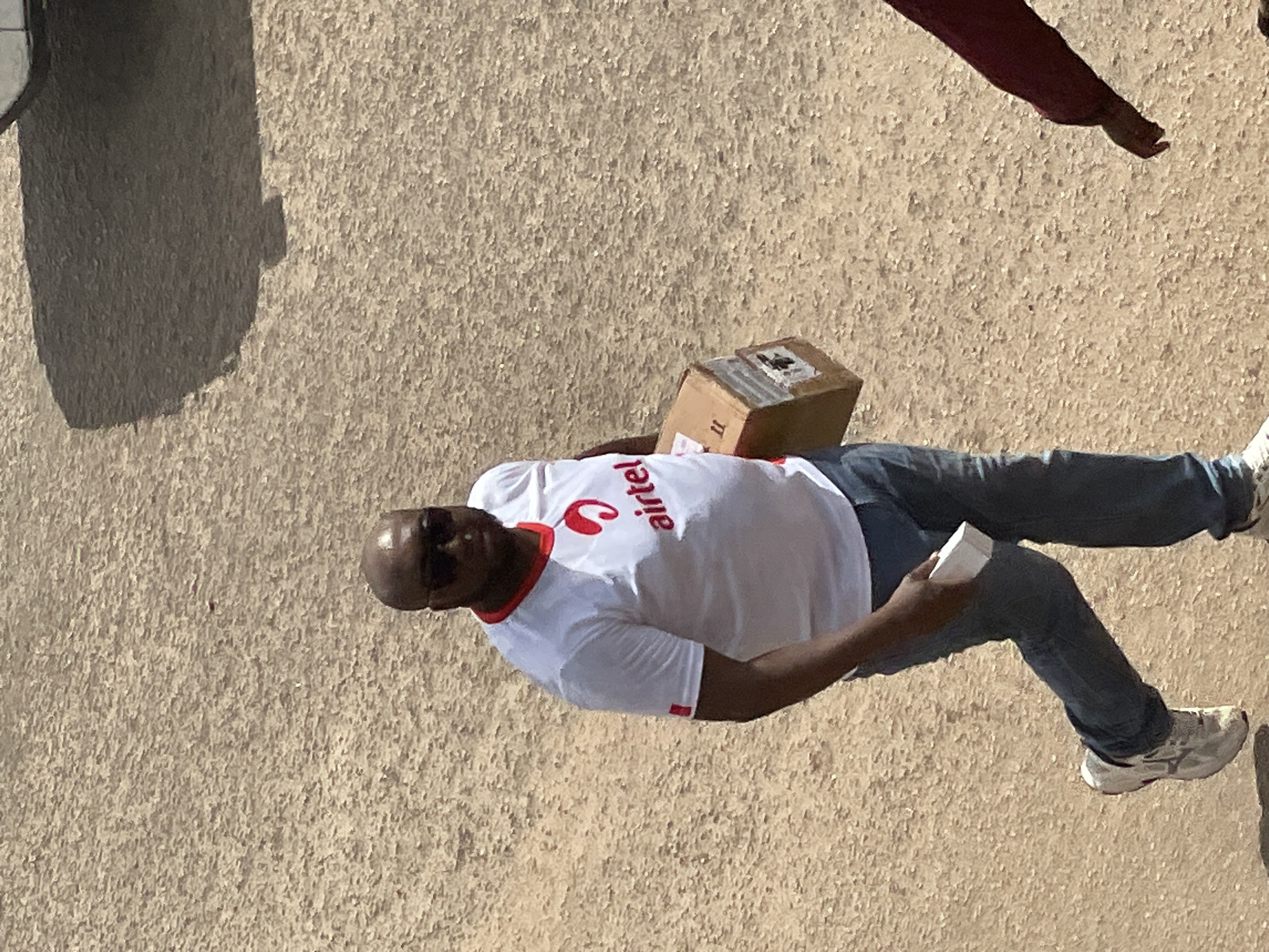 A bald, Black man wearing sunglasses, a white t-shirt with red trim and an "Airtel" logo, and blue jeans walks across a dirt or gravel surface. He is carrying a cardboard box in his right arm and a small white object in his left hand. The man is casting a shadow on the ground to his left. To the right of the frame, a portion of another person's arm wearing a burgundy shirt is visible. The environment appears to be outdoors in a city, possibly a street or pathway.