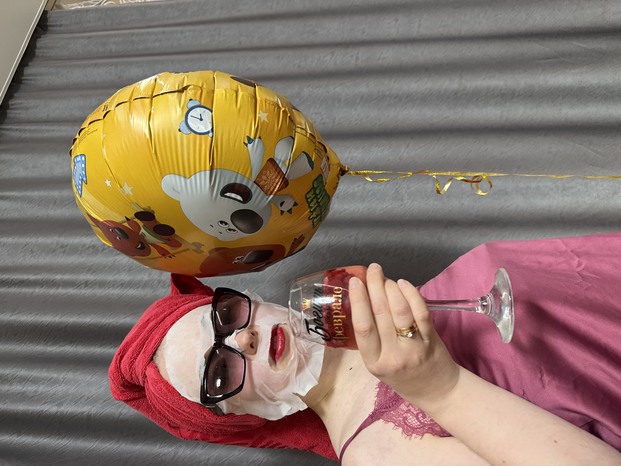A person is depicted from a top-down perspective, lying on their back in what appears to be a bed or couch. They are engaged in a self-care routine, wearing a white sheet mask on their face, large dark sunglasses over the mask, and a red towel wrapped around their head. Red lipstick is visible on their lips. They are dressed in a pink or maroon lace camisole.
The person holds a clear stemmed glass, possibly a wine glass, filled with a reddish beverage, and is taking a sip. On their left ring finger, a gold-colored ring with a small stone is visible. Beside their head, a large, yellow foil balloon floats, adorned with cartoon images of bears, bunnies, clocks, and stars. The balloon appears to be tied with a golden ribbon.
The background consists of a textured gray fabric, likely a bedspread or blanket, and a section of pink fabric. The overall scene suggests a relaxed and indulgent moment, possibly celebrating an event given the balloon.
Visible text on the glass reads "Король Февраля" (King of February) and "ROYAL..." above it. On the yellow balloon, the partially visible Russian text likely says "Днём рождения" (Happy Birthday).