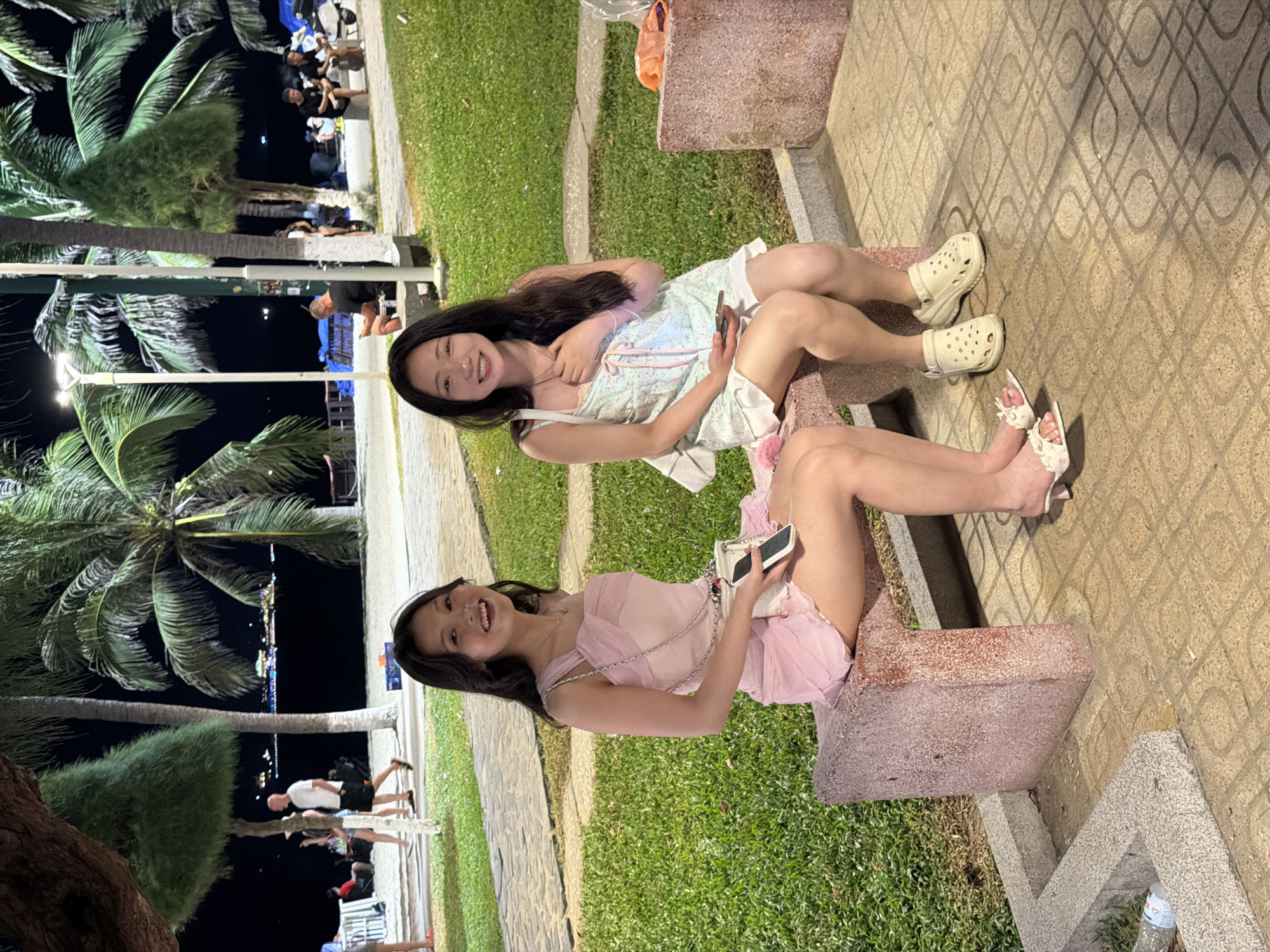 Two young women are sitting on concrete benches next to a sidewalk in Nha Trang, Vietnam. The woman on the left wears a light pink dress and sandals. She holds a smartphone. The woman on the right wears a light blue and white dress and white clogs. Palm trees and a dark, nighttime cityscape are visible in the background. People are walking on the sidewalk. A water bottle is on the ground.