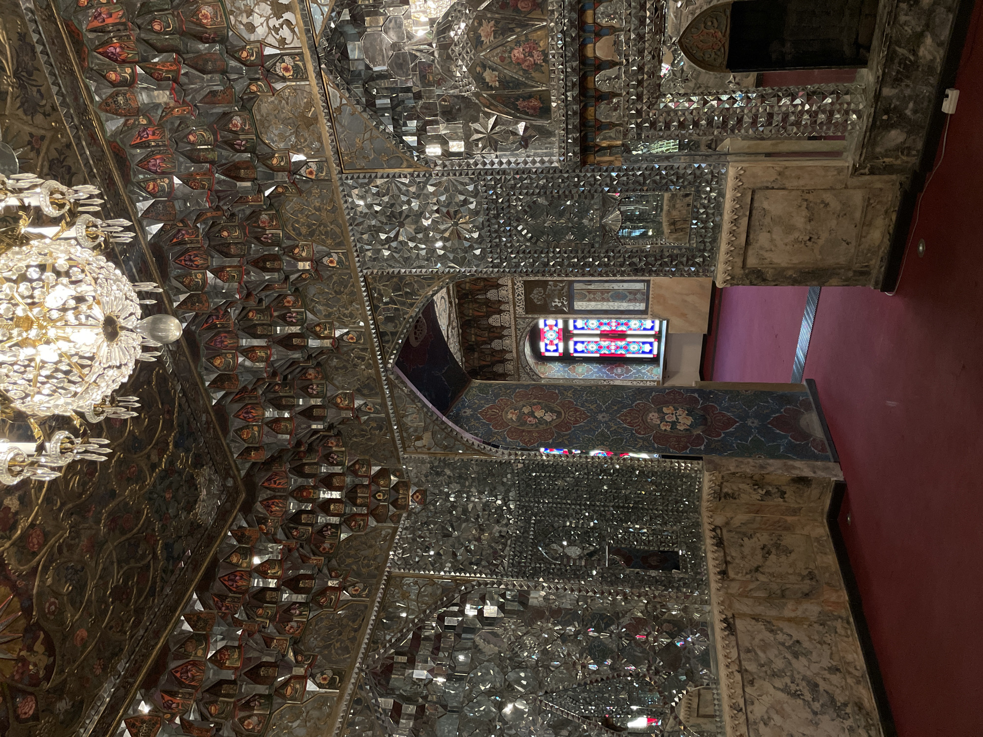 The image showcases the opulent interior of a historical hall or palace in Tbilisi, Georgia, characterized by its elaborate Persian-influenced architecture. The dominant feature is the extensive use of *Ayene-Kari*, a traditional mirror mosaic technique, covering both the walls and ceiling. Thousands of precisely cut mirror pieces are arranged into intricate geometric patterns, creating a dazzling, multi-faceted surface that reflects light.
Interspersed with the shimmering mirror work are richly painted panels and sculptural elements, adorned with intricate floral motifs such as roses and other blossoms, rendered in vibrant reds, oranges, greens, and golds. These painted sections are seamlessly integrated into the overall design, with some protruding to add three-dimensional depth.
On the left, a large, multi-tiered crystal chandelier with illuminated bulbs casts a warm glow, its facets contributing to the sparkling ambiance. Towards the right, a rectangular stained-glass window is visible, featuring a geometric pattern in vivid red, blue, and white glass. Below it, an archway or passage displays additional painted decoration. Portions of the lower walls and architectural frames appear to be made of carved stone or marble with an aged patina. A plain red carpet covers the floor, providing a striking contrast to the highly ornate vertical surfaces.
The scene is devoid of people, allowing the intricate artistry and historical craftsmanship to be the sole focus. A small, modern white electrical outlet is visible on the red wall on the far right. The overall impression is one of grandeur, luxury, and detailed historical design, typical of ceremonial spaces from a bygone era in the region.