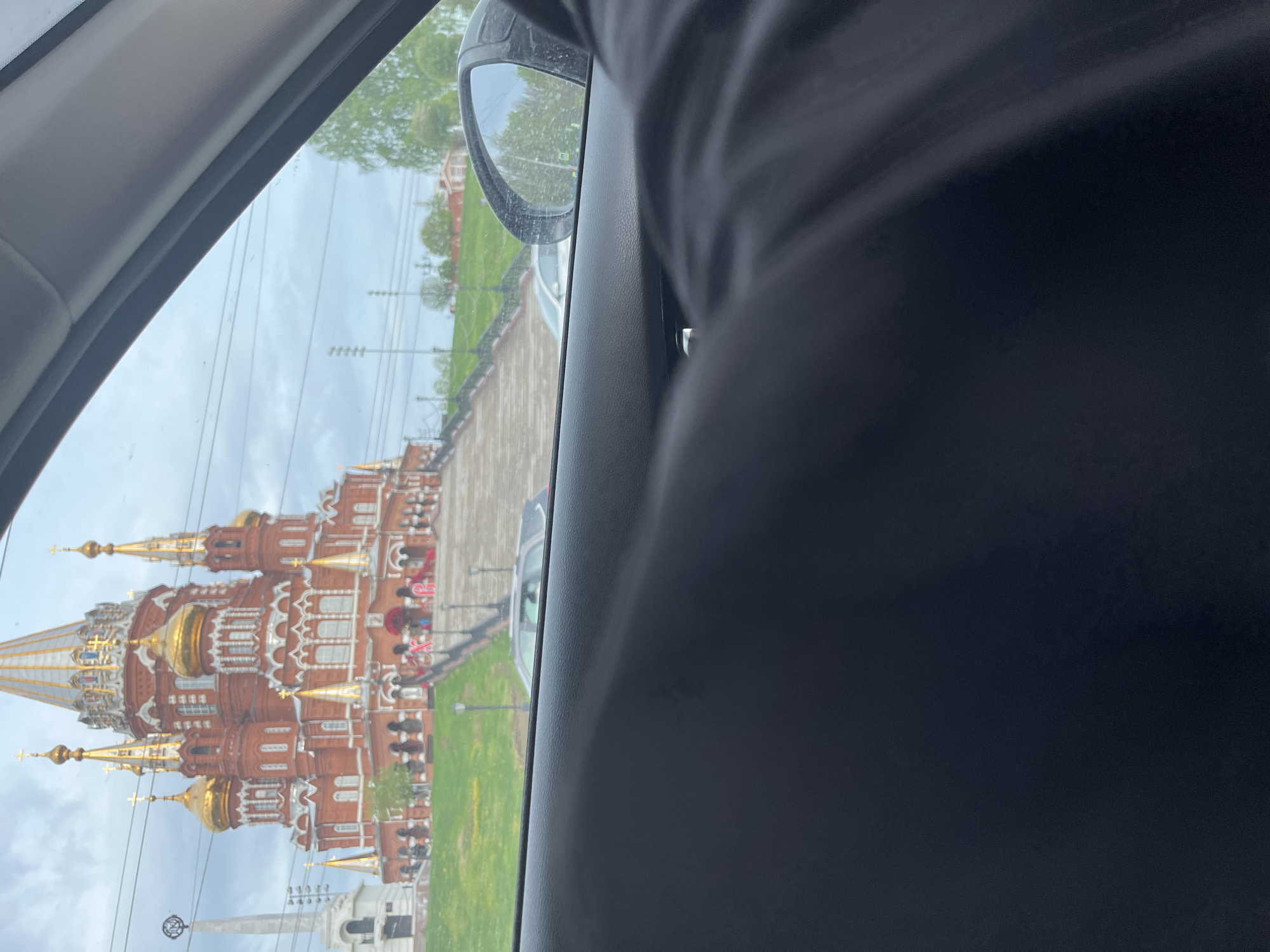 The image shows a view from inside a car, looking out the side window. Reflected in the glass and visible through the window, a large, ornate, reddish-brown building with multiple golden domes and spires is visible. This is likely Saint Michael's Cathedral in Izhevsk, Russia. A green lawn and a pedestrian walkway are visible in front of the cathedral. In the foreground, parts of the interior of the car are visible, including the side mirror and part of a dark seat or clothing.