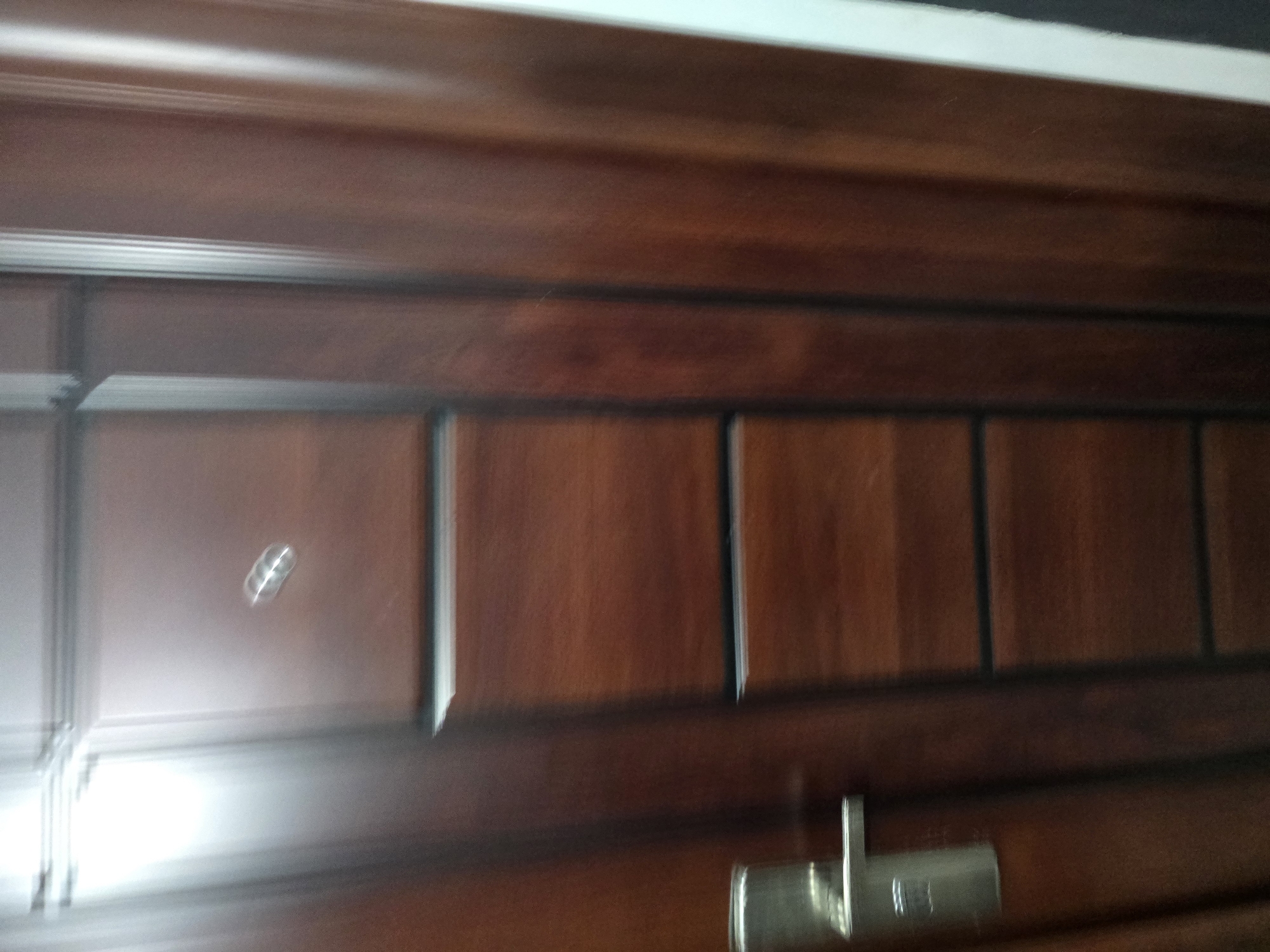 A close-up, slightly blurred shot shows a portion of a dark brown wooden door. The door has a modern design with several horizontal panel divisions. On the left side, a silver-colored door handle is visible. Near the top of the door, a small, metallic peep-hole is present. The lighting is somewhat bright, coming from the left, creating a glare on the upper left section of the door. The image appears to be taken indoors, possibly in a residential building or office. Without further context, it is not possible to determine the specific location within Yambol, Bulgaria.