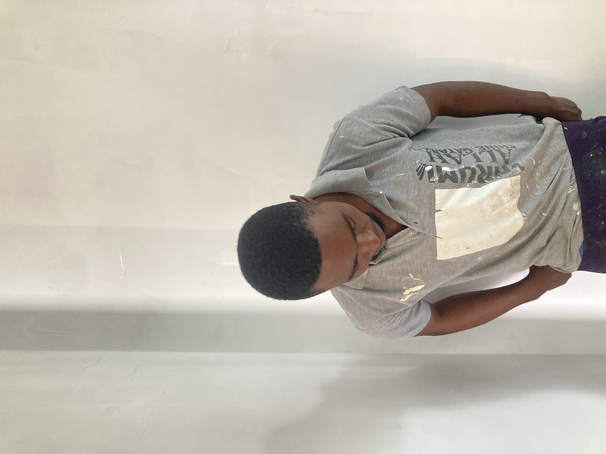 A man is captured from an overhead angle, standing close to a wall. He is wearing a light gray T-shirt with markings on it, along with dark-colored pants. The wall behind him is off-white with slight surface variations. The location, Jos, Nigeria, is not discernible from the image.
