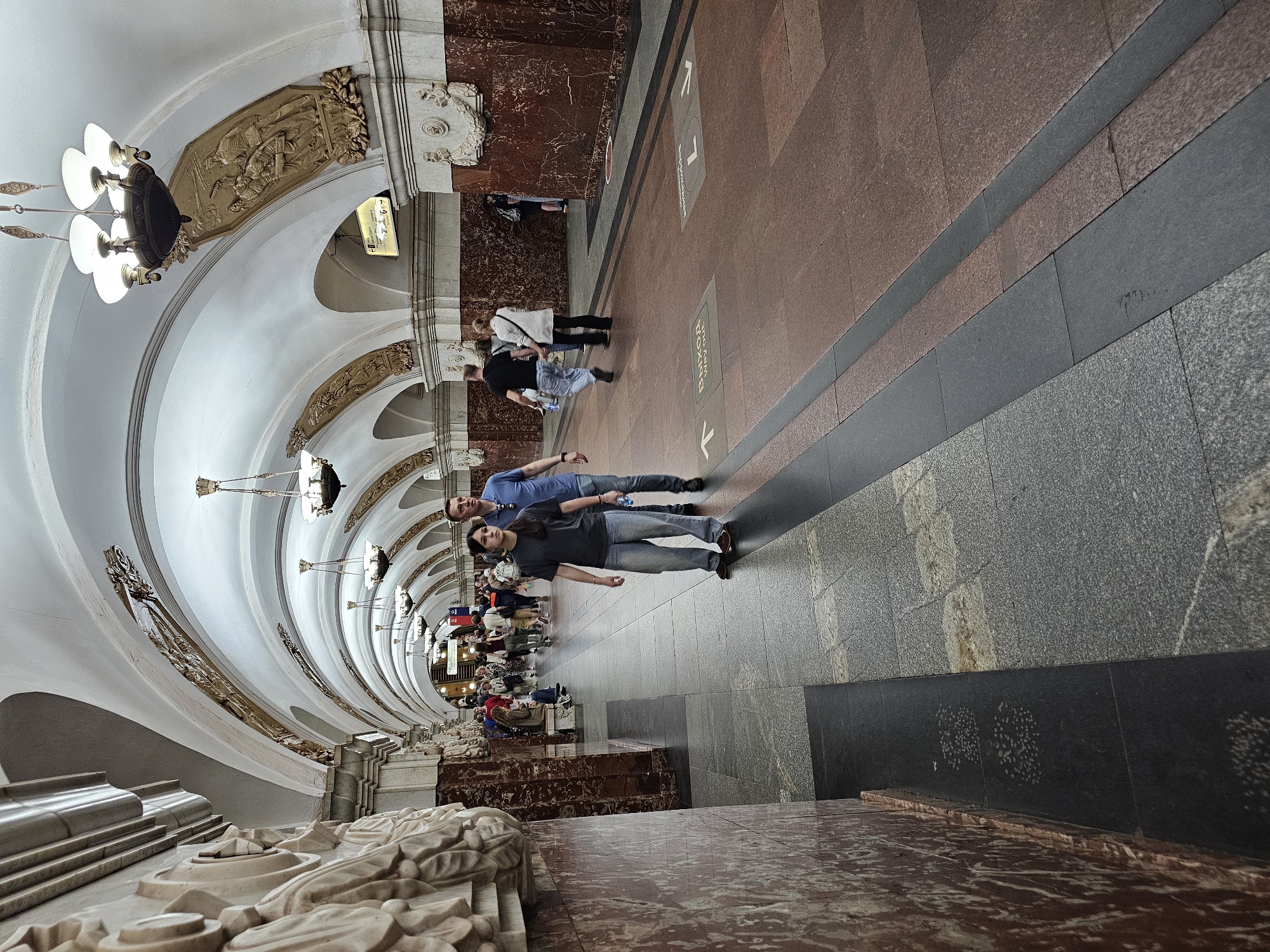 The image depicts a subway station in Moscow, Russia. The station features a curved, ornate ceiling with sculpted details and chandeliers. The walls are covered in a combination of marble and granite panels. Two people stand on the platform, dressed in casual clothing. Several other people are visible in the background, waiting or walking on the platform. Signage is present on the platform floor.