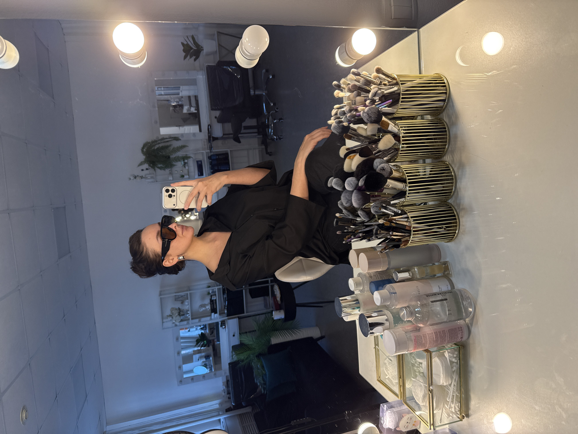 A woman is depicted in a selfie taken in a large, illuminated vanity mirror in Chaikovsky, Russia. She is seated, wearing a black collared shirt and large, dark rectangular sunglasses, with her dark hair styled in an updo and silver stud earrings visible. Her left hand, holding a light-colored smartphone, is raised to capture her reflection. The mirror is framed by multiple exposed incandescent globe lights.
On the white, reflective vanity table in the foreground are several items: two clear glass boxes with gold frames containing cotton pads and cotton swabs, an assortment of skincare and cosmetic bottles including micellar water and toners, and four gold-colored, slatted cylindrical holders filled with numerous makeup brushes of various sizes and types.
The reflection in the mirror reveals a professional interior space. To the left, a dark sofa with a teal throw pillow, a tall white vase holding a green plant, and a small dark side table with a mug are visible. In the center-right reflection, there is a white shelving unit displaying decorative items, another vanity station with a circular mirror and embedded lights, a dark office chair on wheels, and a potted plant. The ceiling is composed of white acoustic tiles with visible light fixtures.