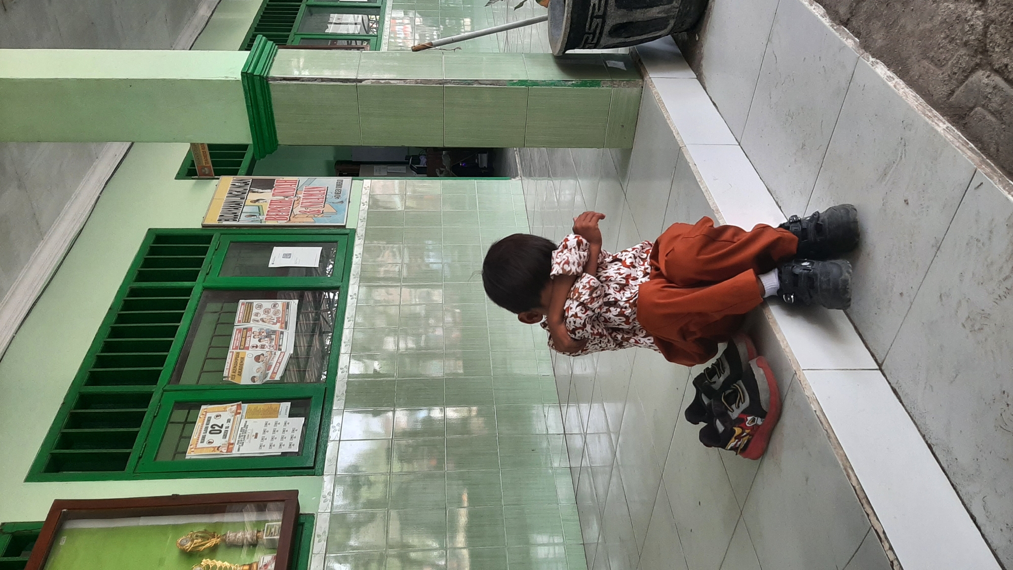 A young child sits on a tiled ledge outside a building. The child is wearing an orange uniform with a patterned shirt and dark pants, and their arms are wrapped around their head, suggesting they might be crying or upset. A pair of black shoes with red soles are placed to their left, and another pair of black shoes are on their feet. The building appears to be a school, with green-painted window frames and walls tiled in light green. Posters are visible on the wall through the windows, and a sign reading "BUDAYAKAN LINGKUNGAN BERSIH INDAH & NYAMAN" (Culture of a Clean, Beautiful, and Comfortable Environment) hangs on the right side of the building. A large green pillar stands to the right of the child. The environment suggests a location in Sumberjo, Indonesia, likely a school.