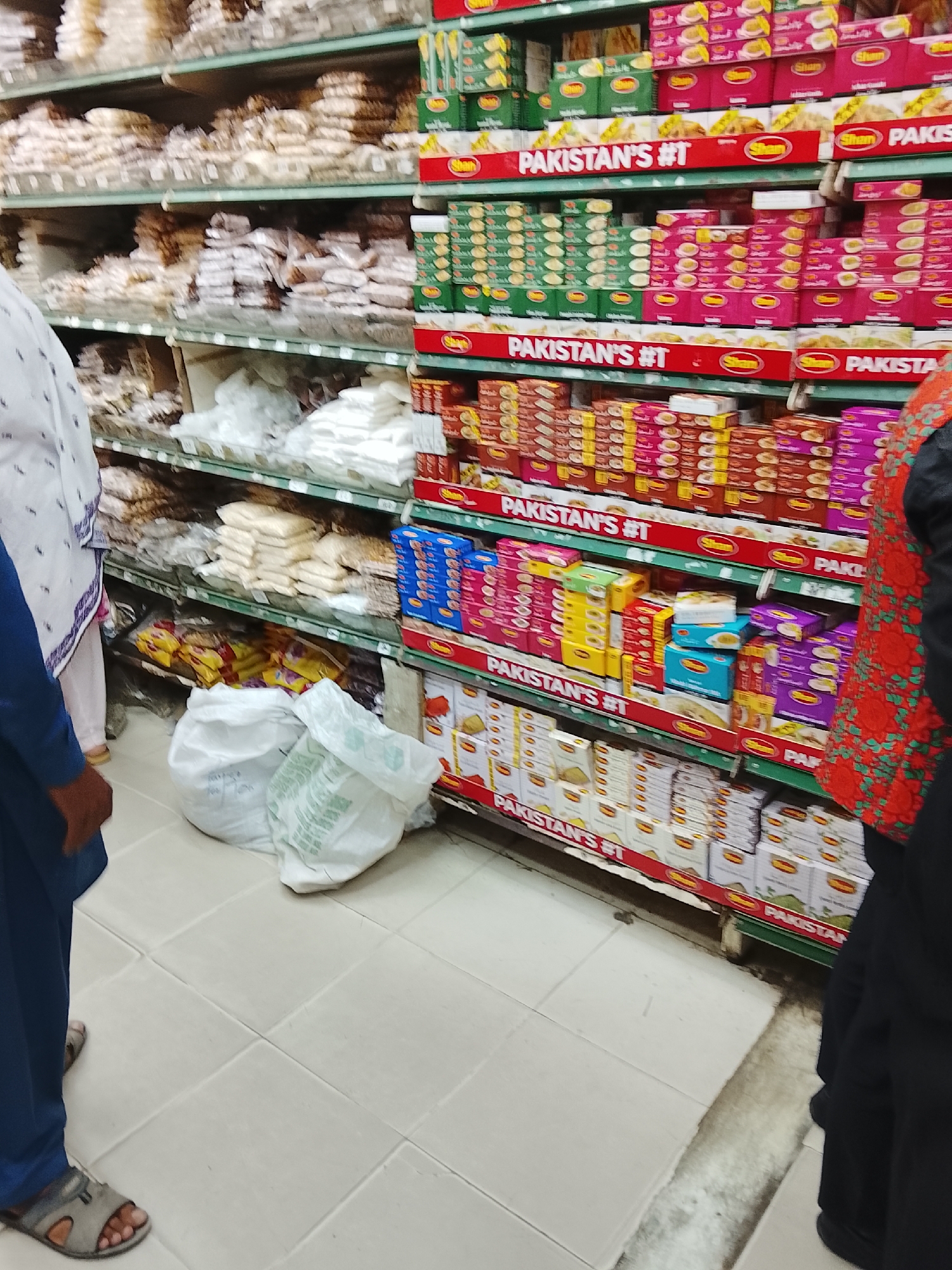 The image captures an indoor scene within what appears to be a grocery store in Gulgasht Colony, Pakistan. Shelves are stocked with various food products, primarily from the brand "Shan," prominently labeled as "PAKISTAN'S #1". The shelves also contain other food items in bulk. Two individuals are partially visible: one wearing a white and purple patterned garment with blue sleeves, and another in black attire with a red patterned shawl. Two white shopping bags sit on the floor in front of the shelves. The floor is tiled.