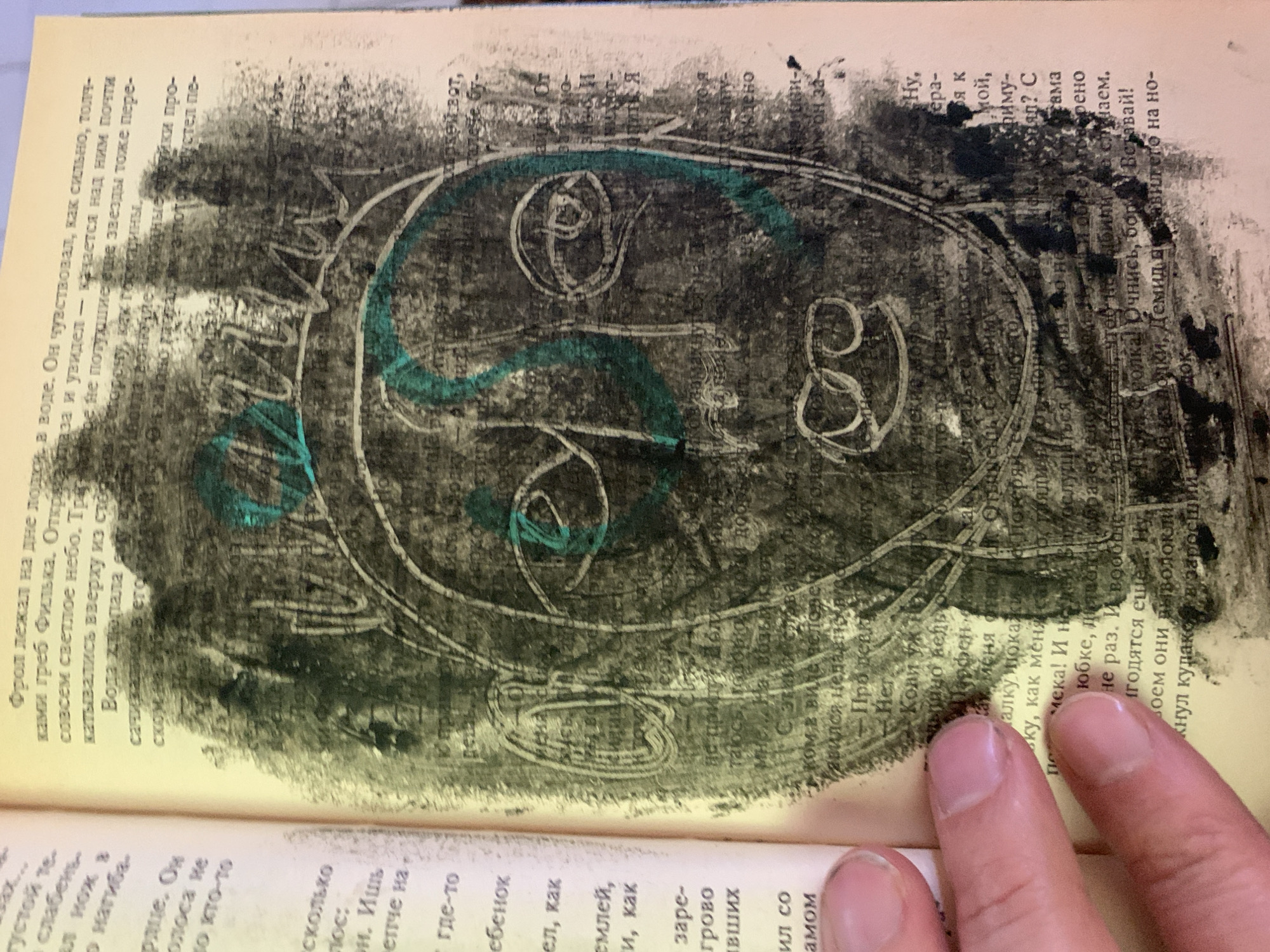 This is a close-up, eye-level, outdoor photograph of a page from a book that has been defaced with drawings. The book page is yellowed and textured. The text is in Russian and is partially obscured by the drawings and a human hand. The drawings are abstract and appear to be a stylized face, rendered in black and green crayon. The face has large, circular eyes with lines inside, a large nose, and a mouth with circles. The head is outlined with a green crayon, and there is a green zigzag line through the center of the face. A human hand with short, clean fingernails is visible in the bottom left corner of the image, holding the book open. The background of the drawing is textured and appears to be made with a rough, smudged black material, possibly charcoal or a dark crayon. The image does not contain any visual elements that would allow for the determination of a location in Ganino, Russia.