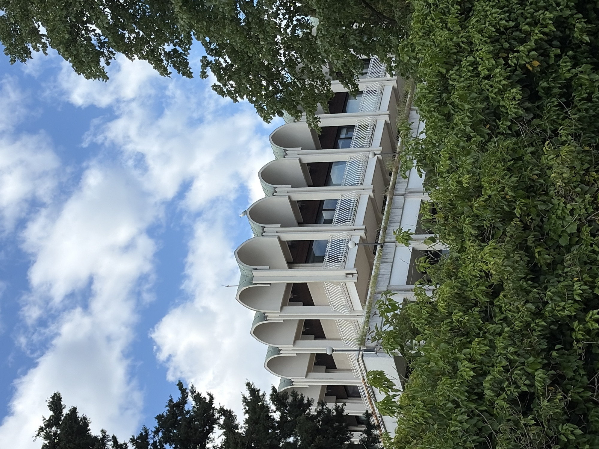 The image depicts a white building with multiple balconies, each featuring a distinct arched design. The sky, dotted with white clouds, is partially visible behind the building. The building is framed by green foliage from trees and bushes on both sides and below. The location is Herceg Novi, Montenegro.