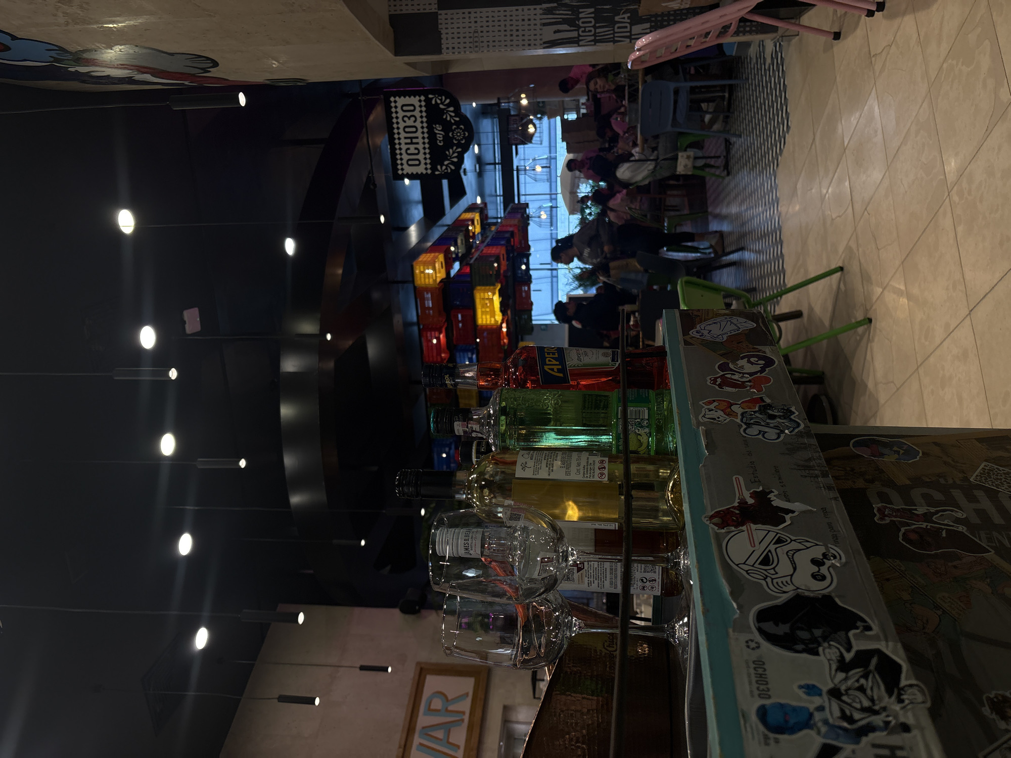 The image depicts the interior of the "OCHO30 café" in Puebla, Mexico. In the foreground, a bar area is visible with a display of liquor bottles and wine glasses. A sign with the name of the café, "OCHO30 café," is displayed prominently. In the background, the café appears busy with patrons seated at tables and interacting. The setting suggests a vibrant, urban environment within a commercial establishment.