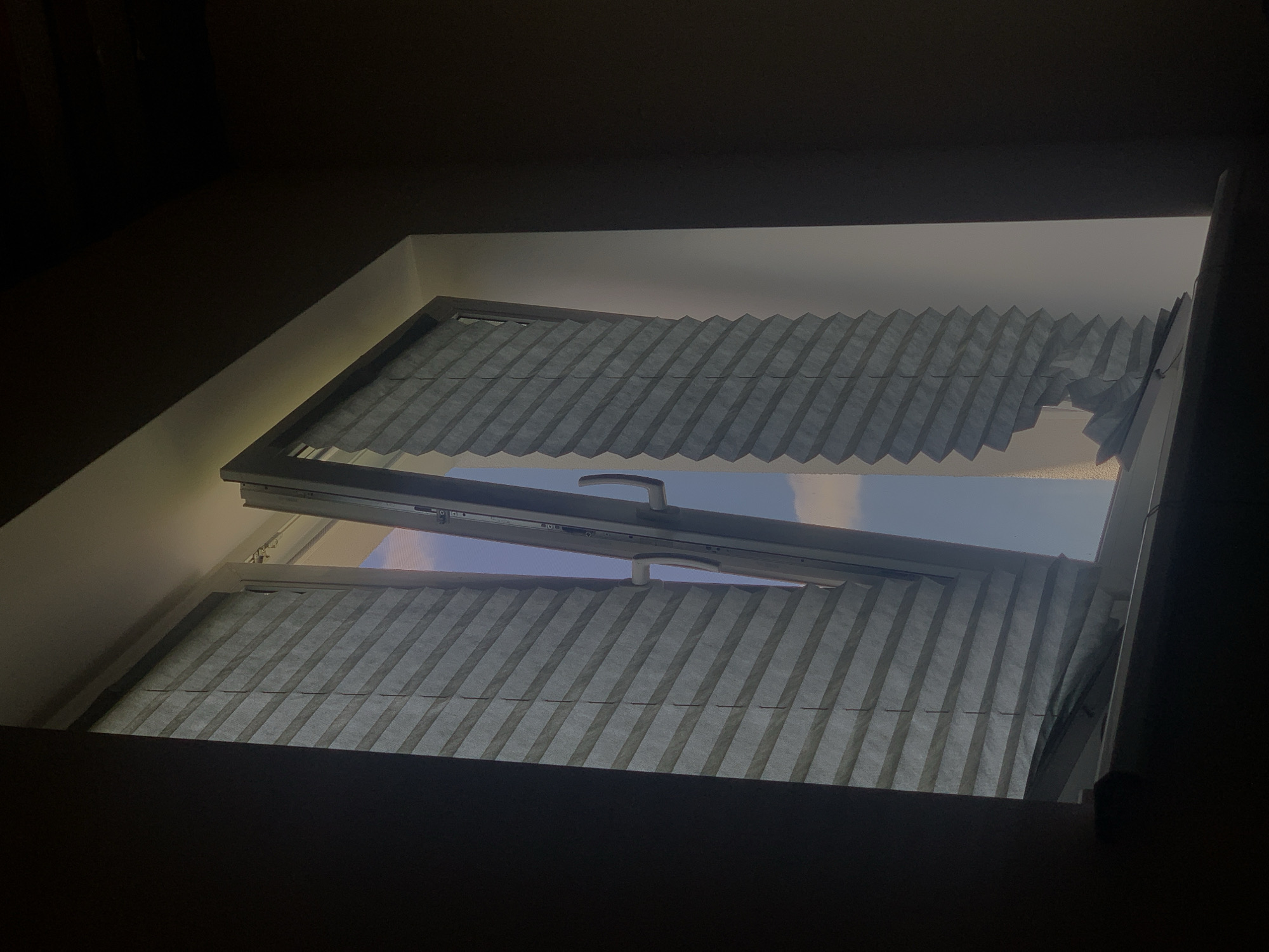 The image depicts a partially opened window with pleated blinds. One side of the window is mostly closed, with the blinds obscuring the view. The other side is open, revealing a glimpse of a blue sky with some clouds. The window frame is light-colored, and a white handle is visible on the open section. The interior of the room appears dark, with shadows cast on the walls and windowsill. The scene is indoors, suggesting a residential or office building. There are no people or animals visible in the image. The location is identified as Minsk, Belarus.
