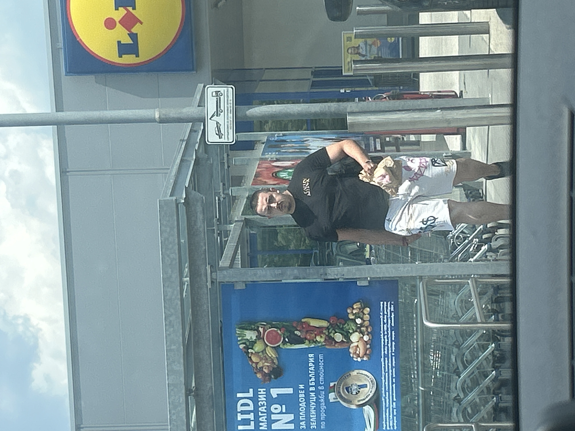 The image shows a man walking in front of a Lidl supermarket in Plovdiv, Bulgaria. The man is wearing a black t-shirt and white shorts with dollar signs printed on them. He is carrying a bag of groceries in his right hand. The Lidl store has its logo visible, a yellow circle with a red dot and blue letters. There is also a large poster advertising the store. The man is walking past a row of shopping carts. The sky is cloudy.