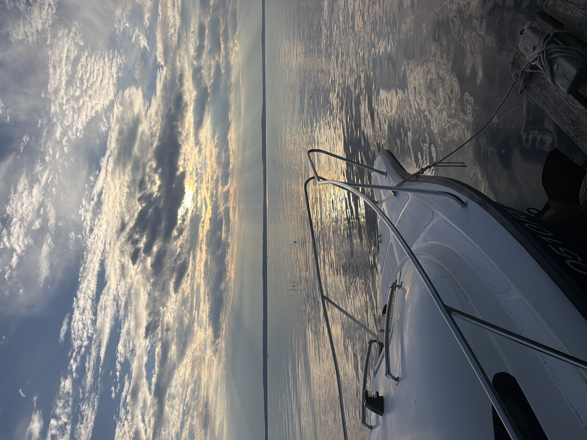 The image captures a serene water scene at sunset, presumably in Stepanovo, Russia. In the foreground, the bow of a white boat is visible, moored to a wooden dock with a rope. The boat's railing gleams under the soft light. The water's surface is calm, reflecting the dramatic sky. The sky is filled with a tapestry of clouds, with the setting sun peeking through, casting rays of light across the water. The distant horizon is marked by a dark, treeline. No people are present in the frame. The overall atmosphere is peaceful and tranquil.