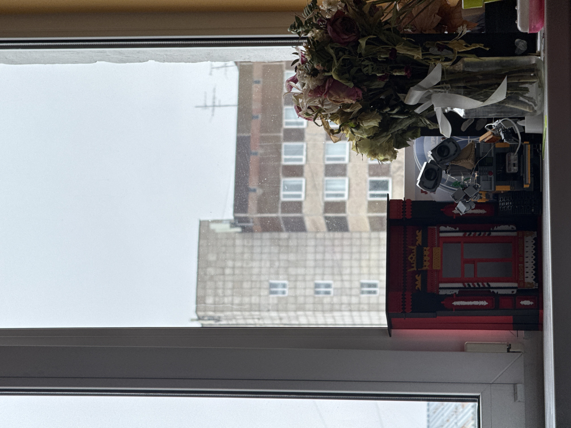 A still-life scene on a windowsill in Perm, Russia. An open window with a white frame reveals a blurred view of a multistory building across the street under overcast skies. The windowsill is cluttered with objects, including a bouquet of dried flowers, a glass jar, electronic components and a structure that resembles a Japanese building crafted from red and black materials. A few dust spots are visible on the window.