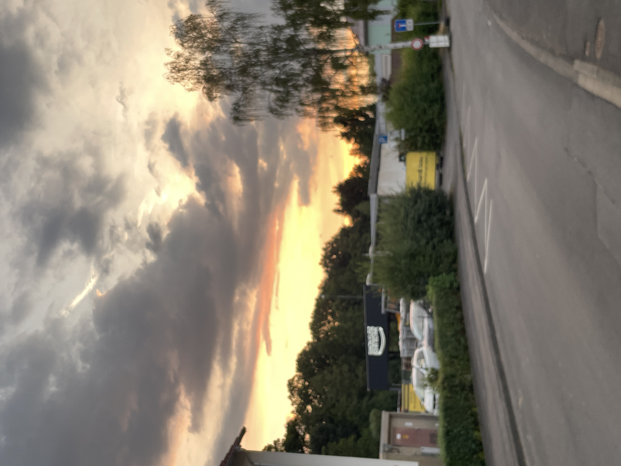 The image depicts a street scene at dusk in Neukirchen, Germany. The sky is filled with dramatic clouds, illuminated by the setting sun which casts an orange and yellow glow. In the foreground, a paved road with white lane markings curves to the right. A sidewalk runs along the left side of the road, bordered by a green hedge. Beyond the hedge, a building with a brown door is visible on the far left, followed by parked cars, including a white sedan and a dark-colored car, and more hedges. Further back, a long, dark-colored building with a prominent white logo featuring text is situated behind trees. A tall, leafy tree with delicate branches stands to the right of this building, its leaves catching the sunset light. Road signs are present on the right side of the image, indicating traffic regulations. The overall impression is one of an urban environment bathed in the warm, fading light of sunset.