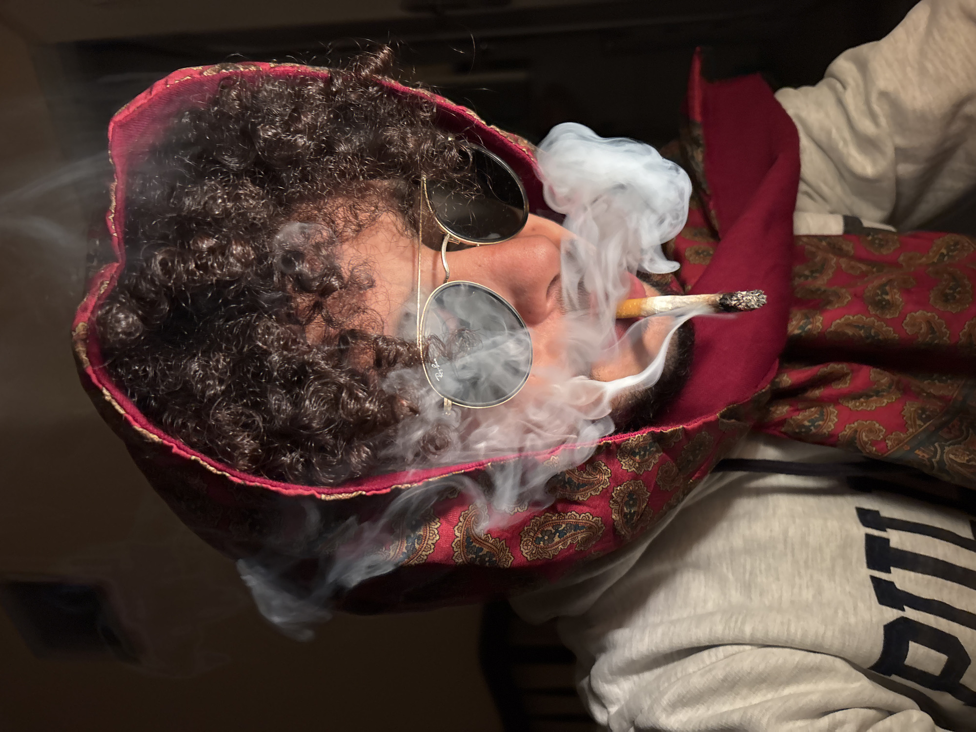 A man with dark, curly hair and round sunglasses is smoking a cigarette. He is wearing a red and gold patterned hat and a grey hooded sweatshirt. He is lying down on a surface with similar red and gold patterns. Smoke surrounds his face. The location is not discernible from the image.