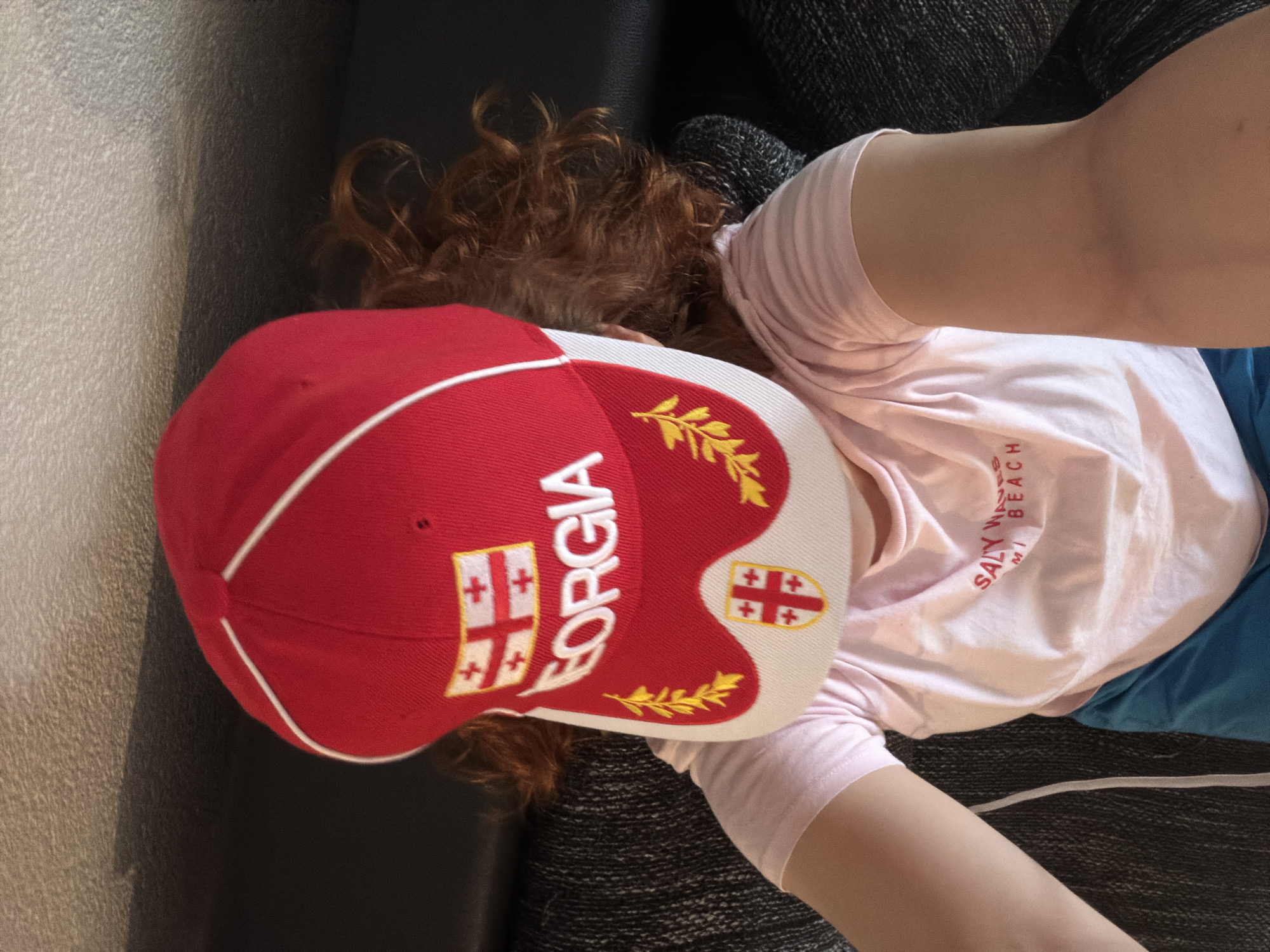 A person wearing a red baseball cap with the word "GEORGIA" and the Georgian flag motif is lying down on a dark, textured surface, possibly a couch or chair. The cap is partially obscuring their face. They are wearing a light pink t-shirt with a graphic that appears to say "SALTY WAVES MI BEACH." The person also appears to be wearing blue shorts. Their reddish-brown hair is visible around the cap. The background is a dark, textured surface. The image is taken from a high angle, looking down at the person. There is no indication of the city of Strasbourg, France in the image.