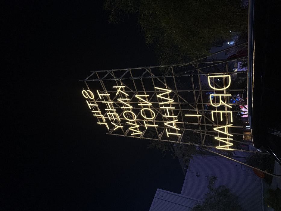 A nighttime scene depicts a large-scale neon light art installation located in Alserkal Avenue, Al Quoz, Dubai, United Arab Emirates. The artwork features yellow neon letters spelling "STILL THEY KNOW NOT WHAT I DREAM," displayed vertically and in reverse. The neon elements are affixed to a multi-tiered, industrial-style metal scaffolding structure that rises prominently against a dark, clear sky. Trees and sparse foliage are visible on both sides of the installation. In the left background, a building with a faint purple-blue illumination is partially visible. The upper portion of a dark vehicle is present in the lower foreground, suggesting the image was captured from a ground-level perspective, likely from within or directly in front of the vehicle. No individuals, specific actions, or interactions are discernible.