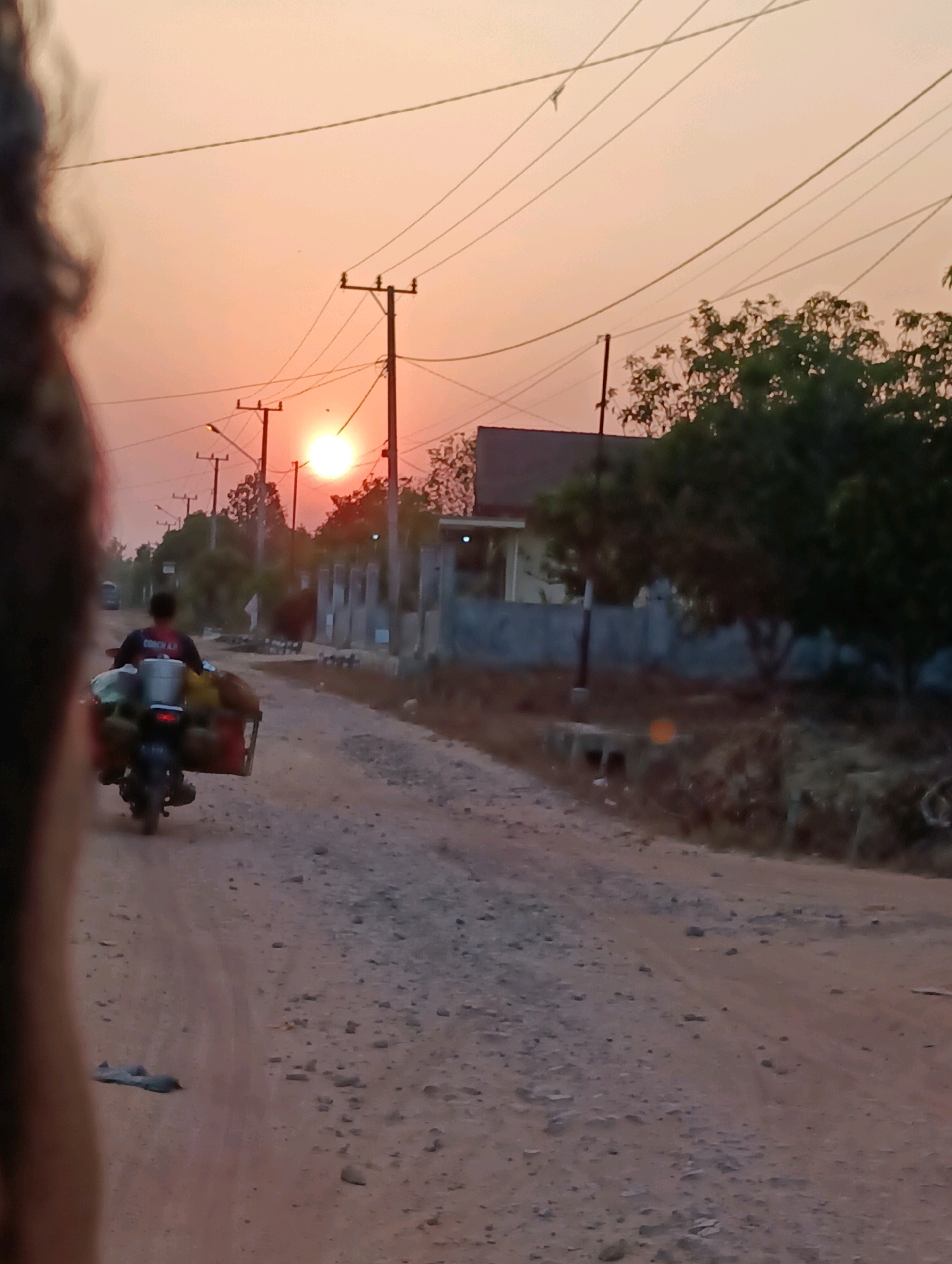 A person rides a motorcycle on a dirt road at sunset in Pematang Jaya, Indonesia. The motorcycle is loaded with goods. The sun is setting in the background, casting a warm glow over the scene. There are utility poles and power lines along the road, and some trees and buildings are visible on either side. The road is unpaved and appears to be made of gravel and dirt. A blurred foreground element, likely a person or vehicle, is partially visible on the left side of the image.