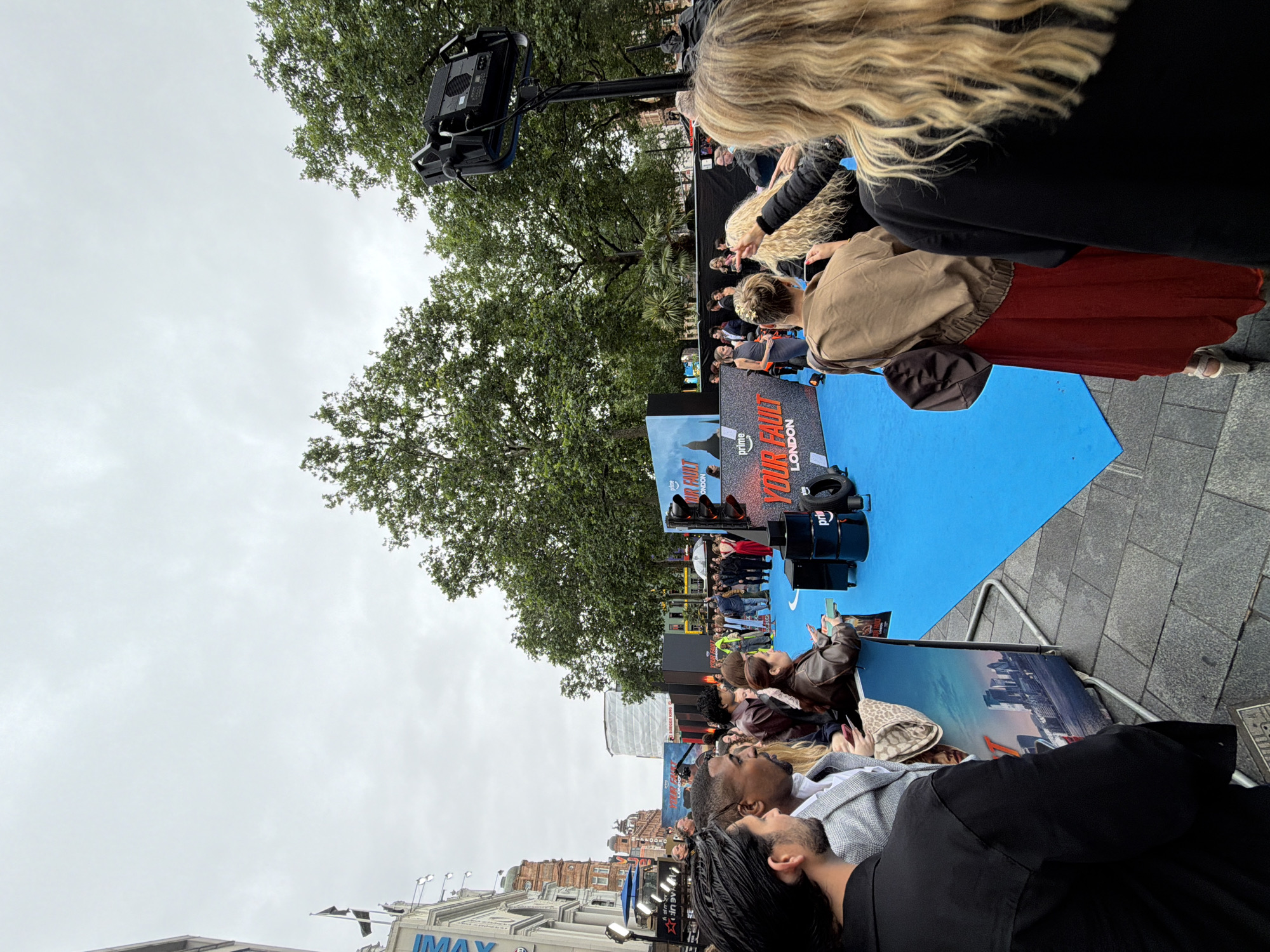 The image shows an outdoor event likely a film premiere or promotional event, taking place on a paved street in the city of London, United Kingdom. The setting appears to be in front of a cinema or large venue, with a building featuring the "IMAX" logo visible on the left. A blue carpet or stage is laid out, with event signage featuring the text "YOUR FAULT LONDON". Several people are present, some standing and watching, while others are more actively involved in the event, possibly as guests or staff. A large lighting rig is visible on a stand in the background. The sky is overcast.