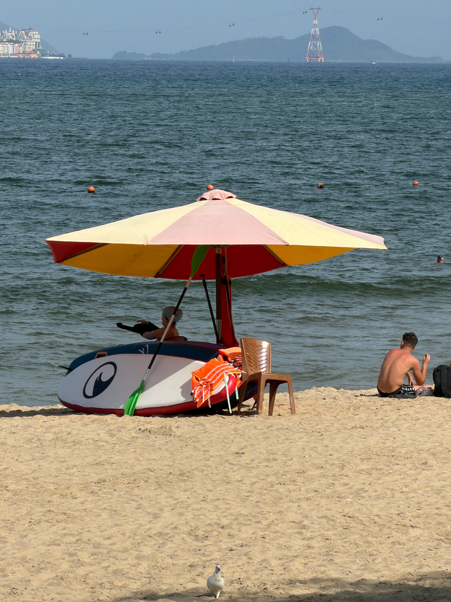 The image depicts a beach scene in Nha Trang, Vietnam, with the ocean in the background and a sandy shore in the foreground. The sea is calm with gentle waves, and a few buoys are visible in the water. In the distance, a resort complex with distinctive architecture and a large electrical tower on a mountain are prominent features of the Nha Trang coastline.
On the beach, a colorful umbrella shades a stand-up paddleboard (SUP) which is resting on the sand. A person, wearing a head covering and possibly a rash guard, is seated on the SUP, partially obscured by the umbrella. Next to the SUP, an orange life vest and a brown plastic chair are placed.
To the right of the umbrella, a man is sitting on the sand, facing away from the camera. He is wearing dark swim trunks and appears to be engaged with something in his lap. A black bag is positioned beside him.
A single bird, likely a seagull, is visible in the foreground on the sand. The overall atmosphere suggests a relaxed day at the beach.