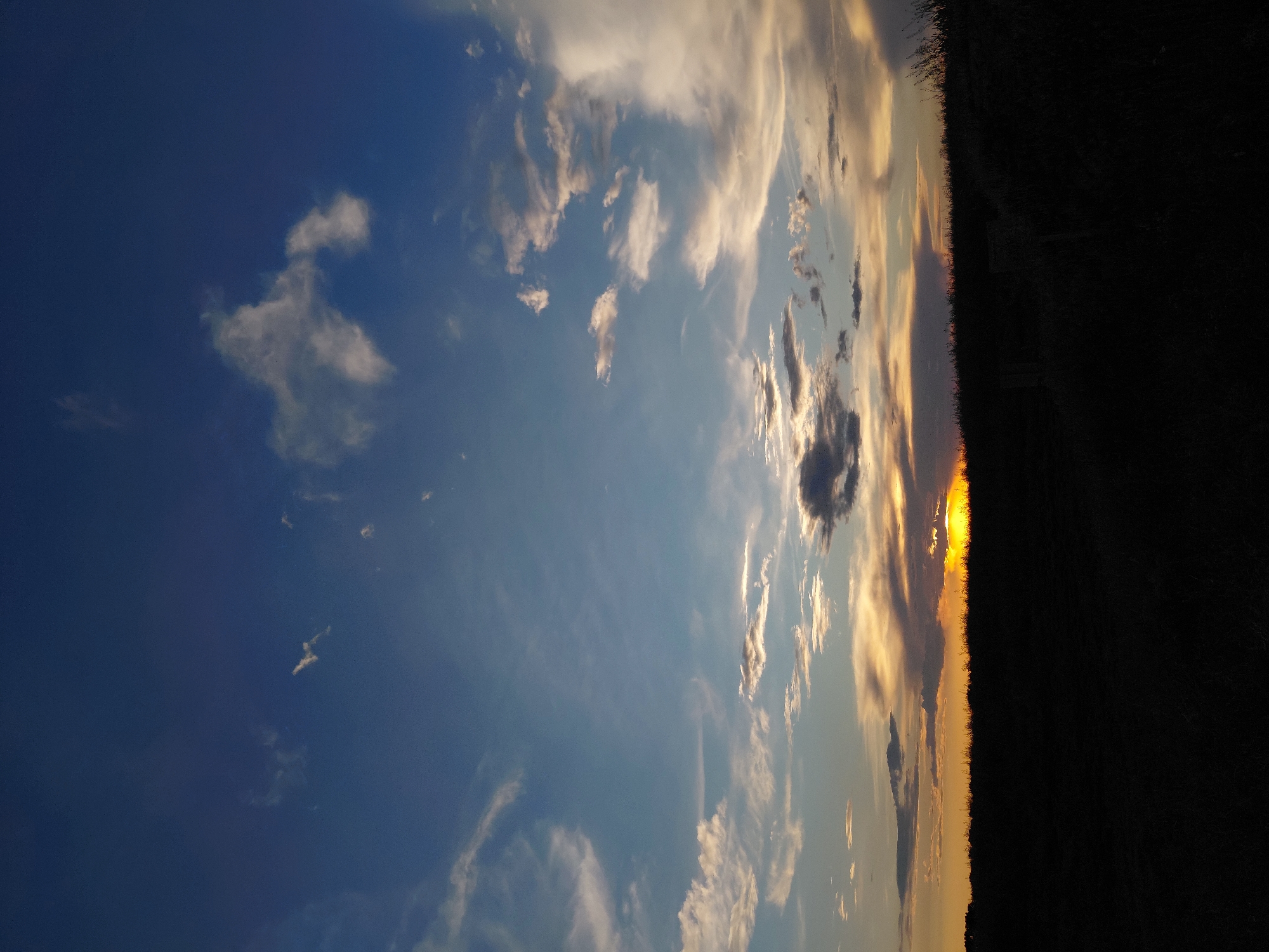 The image captures a sunset scene over a dark, silhouetted landscape, likely a field or hilly terrain. The sky is a gradient of deep blue at the top, transitioning to lighter blues and then to hues of orange and yellow near the horizon where the sun is setting. Scattered clouds, some appearing wispy and others more dense, are illuminated by the setting sun, creating patches of white, gray, and golden light against the darkening sky. The lower portion of the image is completely in shadow, obscuring details of the ground and vegetation. Two faint, upright objects, possibly fence posts or small structures, are barely visible within the dark silhouette on the right side of the image. There are no people or animals visible in the frame. The location is identified as Ustyvytsia, Ukraine, suggesting a rural or natural setting.