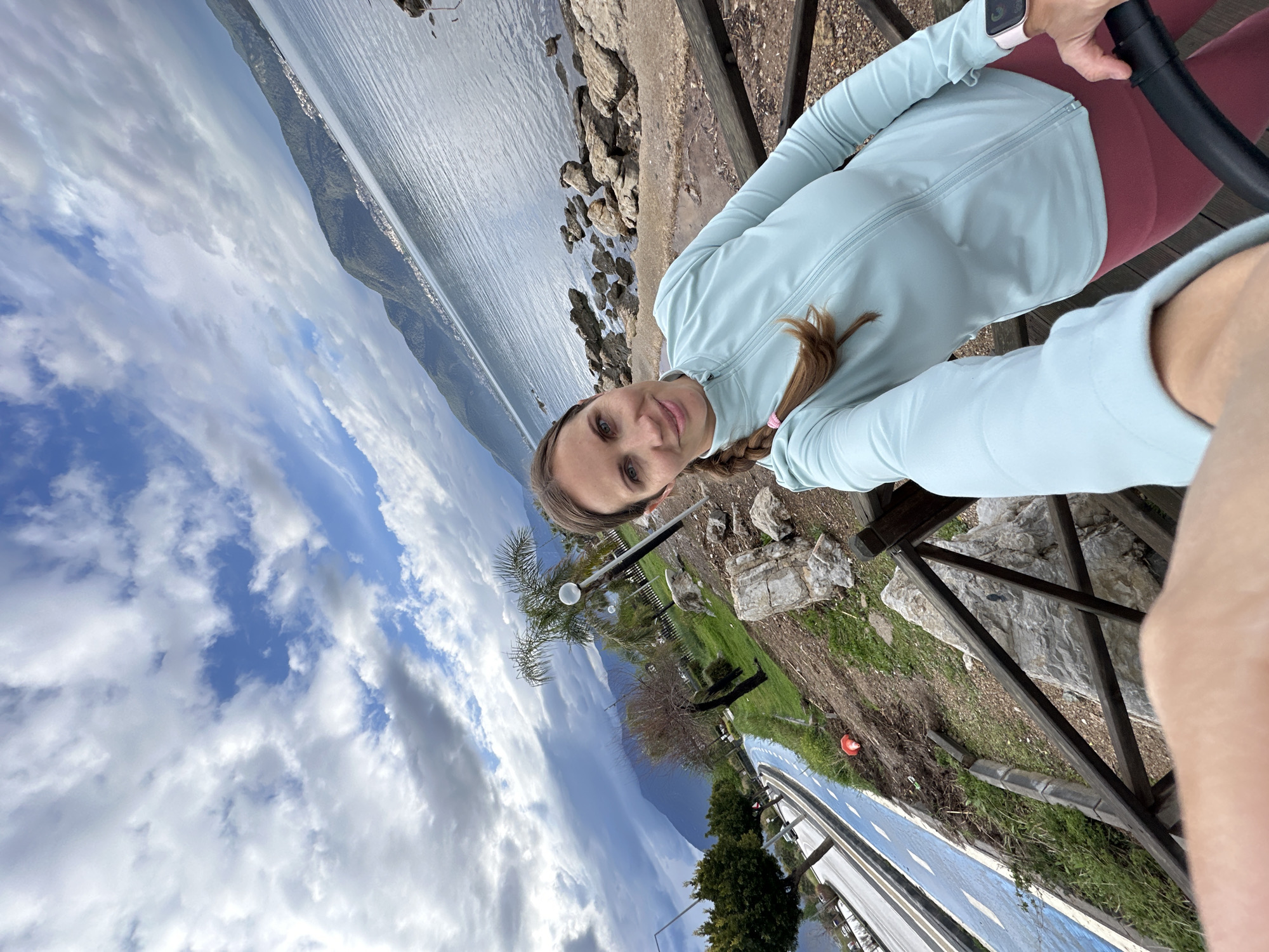 A woman is taking a selfie near a lake in Fethiye, Turkey. She is wearing a light blue jacket and red leggings. She has a braid in her hair and is holding a black handlebar. Behind her, there's a rocky beach and a body of water, possibly the Mediterranean Sea. In the distance, there are mountains. There is also a wooden fence, trees, and a road visible. The sky is cloudy with patches of blue.