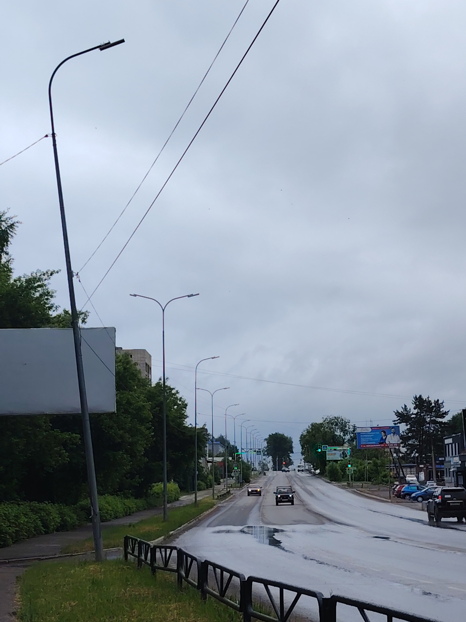 This forensic description details a daytime scene on Vokzalnaya Street in Chaikovsky, Perm Krai, Russia, observed under an overcast sky following recent rainfall.
The perspective is from a pedestrian area on the left side of a multi-lane asphalt road, looking approximately east-northeast up a gentle incline. The road, which is wet with visible puddles, consists of at least four lanes, two for each direction, separated by a solid white line.
In the immediate foreground on the left, a tall, dark streetlamp with a single, curved arm supporting a modern LED light fixture stands prominently. Behind it, a large, blank white rectangular sign is mounted on a pole. A black metal barrier with a repetitive V-shape design runs along the grassy verge.
Further down the left side, a concrete sidewalk is bordered by dense, lush green trees and shrubs. Additional streetlamps, featuring double curved arms, line the road. In the mid-distance, behind the foliage, a multi-story building with a light-colored, possibly concrete, facade is partially visible.
On the right side of the road, several vehicles are parked or stopped. A dark SUV, possibly a Mitsubishi Pajero, is seen from the rear, situated in a parking area. Other parked cars include a blue hatchback, a red sedan, and a light-colored car. Above these, a blue billboard displays images of two men (one younger, one older) and text in Cyrillic, including the address "ул. Вокзальная 43-4" and a phone number. Adjacent to this, a commercial building with "АВТО" (Auto) visible on its facade is partially obscured by trees and the billboard.
Traffic is moving away from the viewer on the uphill road. A dark SUV occupies the left lane, and a lighter-colored sedan is in the right lane. Further up the incline, two additional cars, one light and one dark, are visible. Multiple sets of traffic lights, configured horizontally across the road, display green signals, indicating active traffic flow.
The sky is uniformly overcast and light grey, with several tiny dark specks, likely birds or insects, scattered across it. No people are visible in the scene.