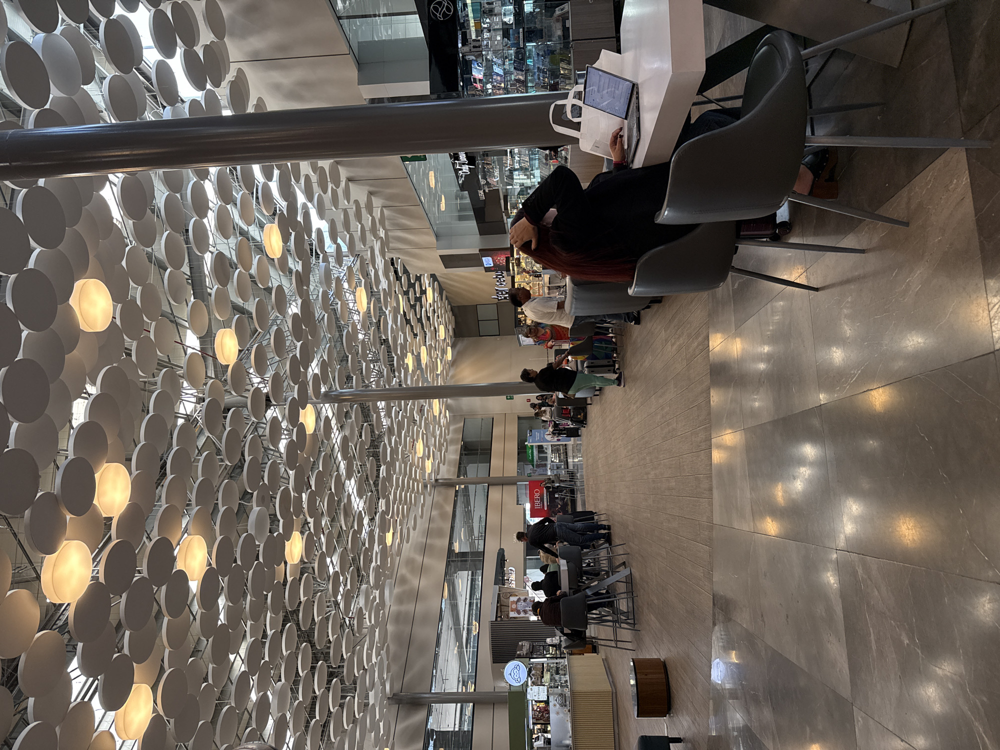This image depicts a bustling concourse within the Puebla International Airport (Aeropuerto Internacional de Puebla, PBC) in Huejotzingo, Puebla, Mexico.
The scene is illuminated by numerous circular, warm-toned light fixtures integrated into a distinctive modern ceiling composed of hundreds of suspended white circular acoustic panels of varying sizes. A large, dark gray cylindrical column is prominently visible on the right. The floor transitions from polished gray marble tiles with subtle white veining in the foreground to light brown wooden planks further into the concourse. Walls are predominantly light-colored, featuring a band of dark-tinted windows above, suggesting a second level.
In the immediate foreground on the right, a person with long, auburn hair, dressed in a dark top, sits facing away from the camera at a white rectangular table with a dark base. They are actively using an open silver laptop displaying a user interface, with two white paper bags placed next to it. The person's left hand is raised to their head, fingers near their hair. Their left foot, wearing a dark shoe with a wooden heel, is visible beneath the table.
In the mid-ground on the left, several individuals are gathered around high-top tables with dark gray stools, consistent with a cafe or waiting area. One person in a dark jacket stands facing the tables. A coffee shop or cafe, identified by a "Bubu" sign, is visible behind them. Further right, a man in a light-colored shirt stands near a woman in a colorful garment, with luggage visible nearby. Another retail space with "Only Luxury" signage and display cases showcasing various products is situated behind these individuals. A red "IBERO PUEBLA" sign is also visible in the background.
Approximately fifteen to twenty people are present throughout the concourse, either seated, standing, or walking. Actions include working on a laptop, standing, conversing, and moving through the space, characteristic of an active airport terminal.