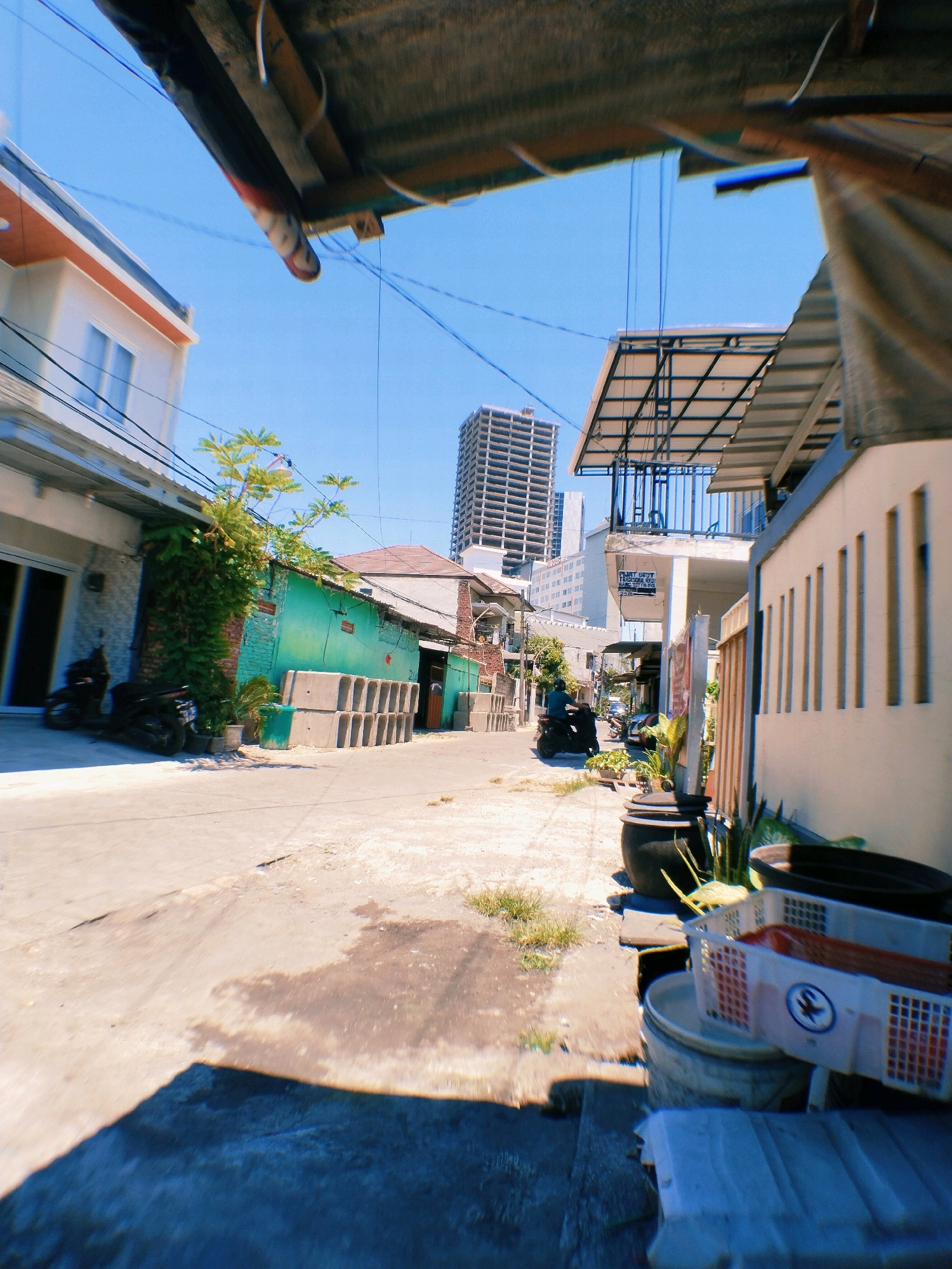The image depicts a street scene in Surabaya, Indonesia. The foreground is dominated by a large shadow cast by an unseen object, possibly a building or structure. To the right, white plastic crates are stacked on top of each other, one of which has a blue and white circular logo. Behind the crates are large, dark cylindrical containers and some greenery.
Moving further down the street, a wall with vertical indentations is visible on the right. Across the street, a vibrant green building with a corrugated metal roof is present, with a stack of concrete blocks lined up in front of it. A motorcycle is parked next to a building on the left, which has white siding and dark framed windows.
In the background, a multi-story building under construction, characteristic of urban development, stands tall against a clear blue sky. A metal awning structure is visible extending over a walkway or entrance. The street itself appears to be paved and shows signs of wear and some dampness. A person is riding a motorcycle away from the camera down the street. The overall impression is of a somewhat industrial or developing urban area.