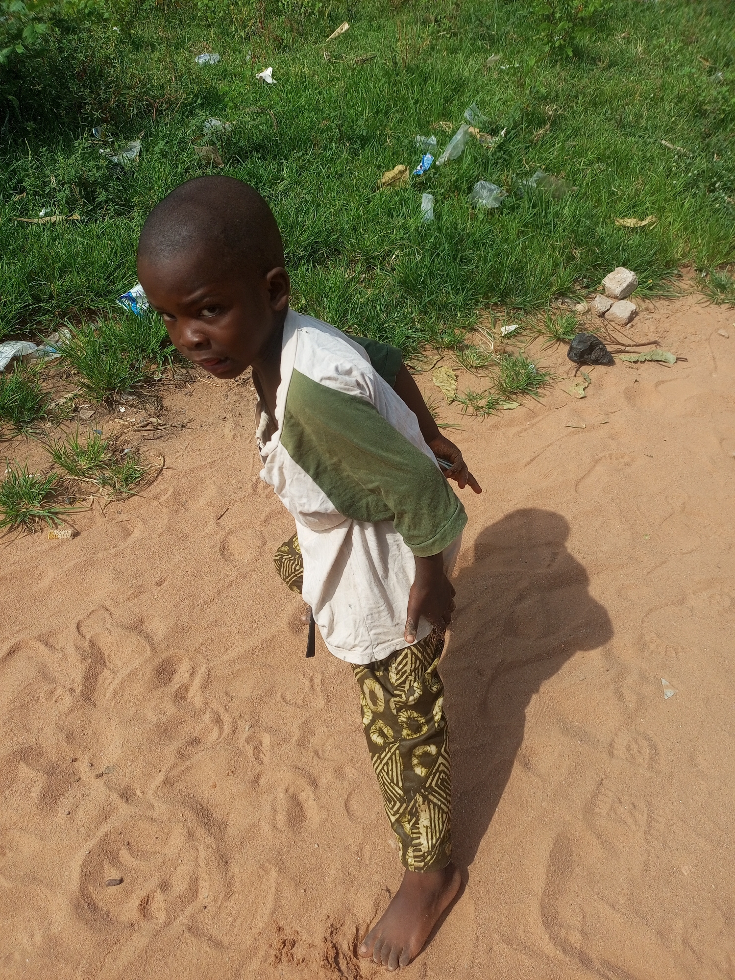 In Tempure, Nigeria, a young boy stands on a sandy surface with a patch of green grass and scattered debris behind him. He is wearing a white shirt with green accents and patterned pants. His right arm is extended, and his gaze is directed slightly upwards and to the left. The lighting suggests a sunny day, casting a shadow to his right. The environment appears to be an outdoor setting with a mixture of natural elements and signs of human presence through the discarded litter.