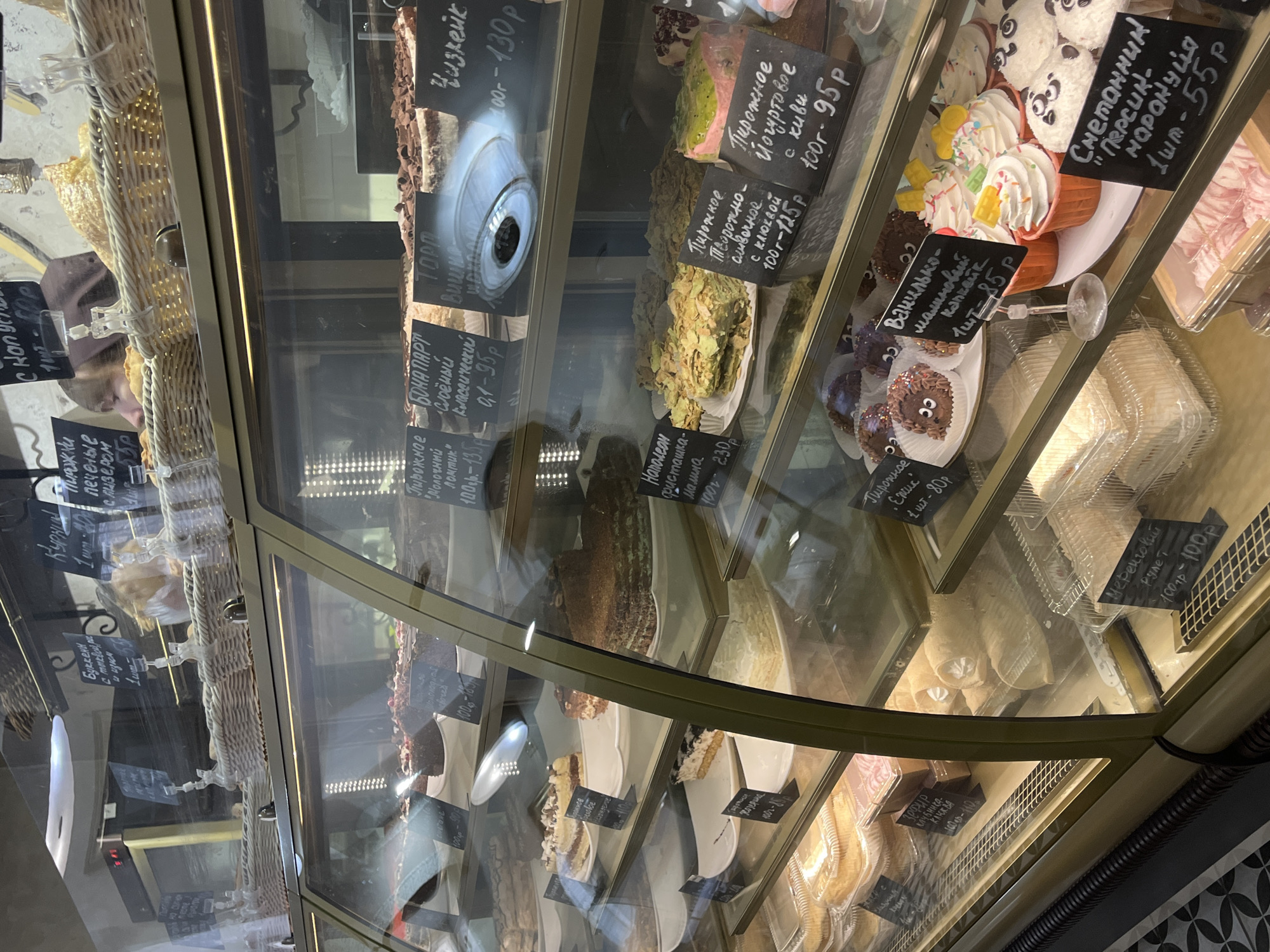 The image displays the interior of a bakery or sweet shop, centered on a multi-tiered glass display case filled with various baked goods and desserts. This scene is located in Neftekamsk, Russia.
The display case features a golden-bronze colored frame and shelves illuminated by internal lighting. Numerous cakes, pastries, and individual sweets are meticulously arranged on white plates, in clear plastic containers, or directly on the shelves. Products include layered cakes, chocolate cakes, cheesecakes, cupcakes with white frosting and sprinkles, cupcakes designed as hedgehogs and pandas, and cream-filled rolls. Each item or section is identified by small black chalkboards displaying names in Russian Cyrillic script and prices in rubles. Examples of visible labels include "Napoleon" (230₽), "Cheesecake" (130₽), "Yogurt Pastry with kiwi" (95₽), and "Sour Cream Cake Peach-Passion Fruit" (55₽).
On the uppermost shelf, several light-colored woven baskets contain additional baked goods. A partial reflection of a person's head and hair is visible in the upper left glass panel of the display case, indicating an individual's presence on the opposite side. The floor visible beneath the display case is covered with black and white patterned tiles. No direct human actions or interactions are depicted in the foreground.