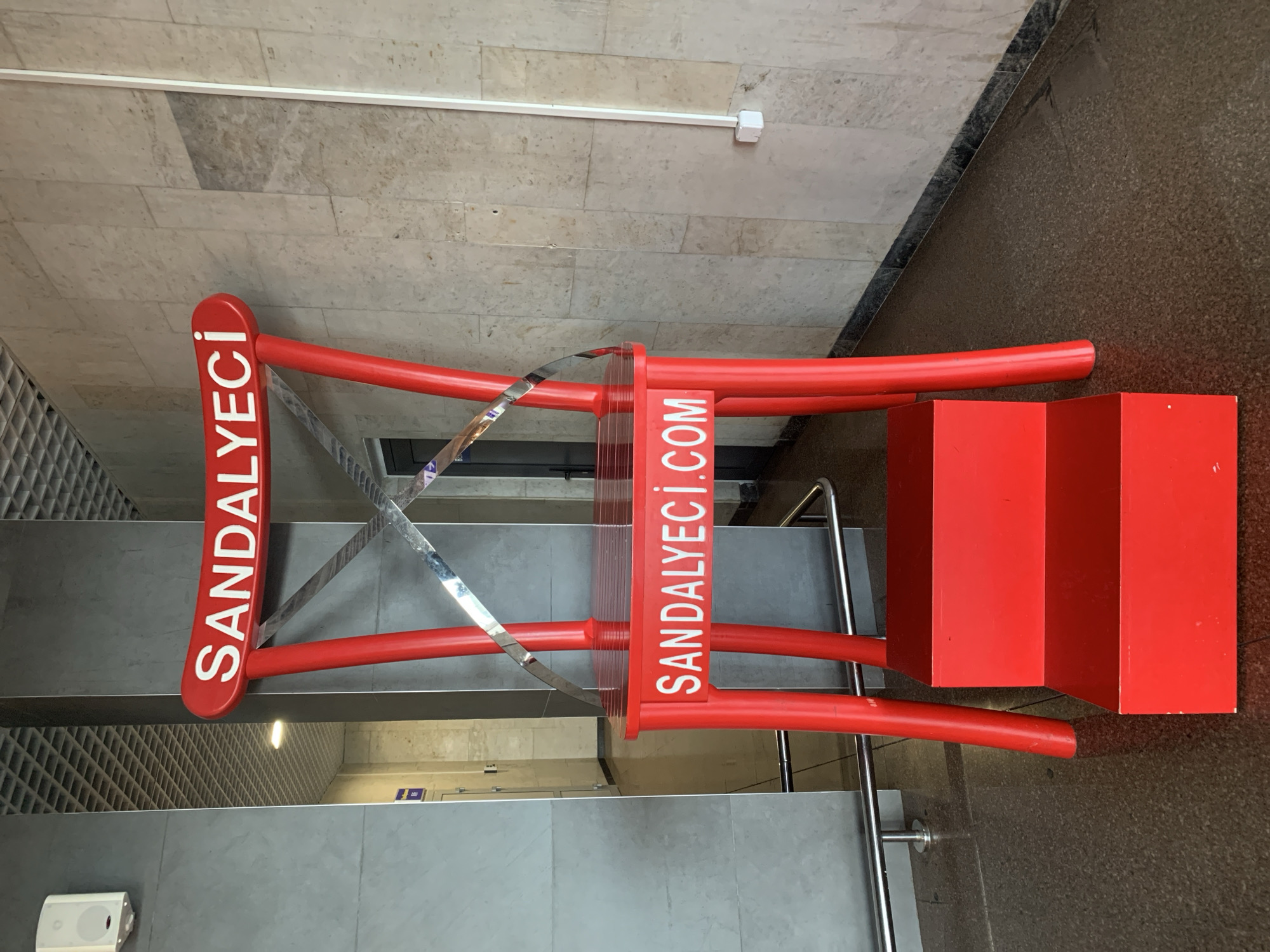 An oversized, bright red chair with the words "SANDALYECI" emblazoned on its back and "SANDALYECI.COM" on its seat stands prominently in an indoor public space. The chair has two steps leading up to its seat, also painted bright red. The flooring appears to be dark speckled granite, and the walls are clad in light-colored stone tiles and grey panels. A white electrical conduit runs vertically up the wall behind the chair. The scene suggests a commercial or advertising display, possibly in a shopping center or transportation hub. While the image was captured in Minsk, Belarus, this information is not visually evident from the photograph itself.