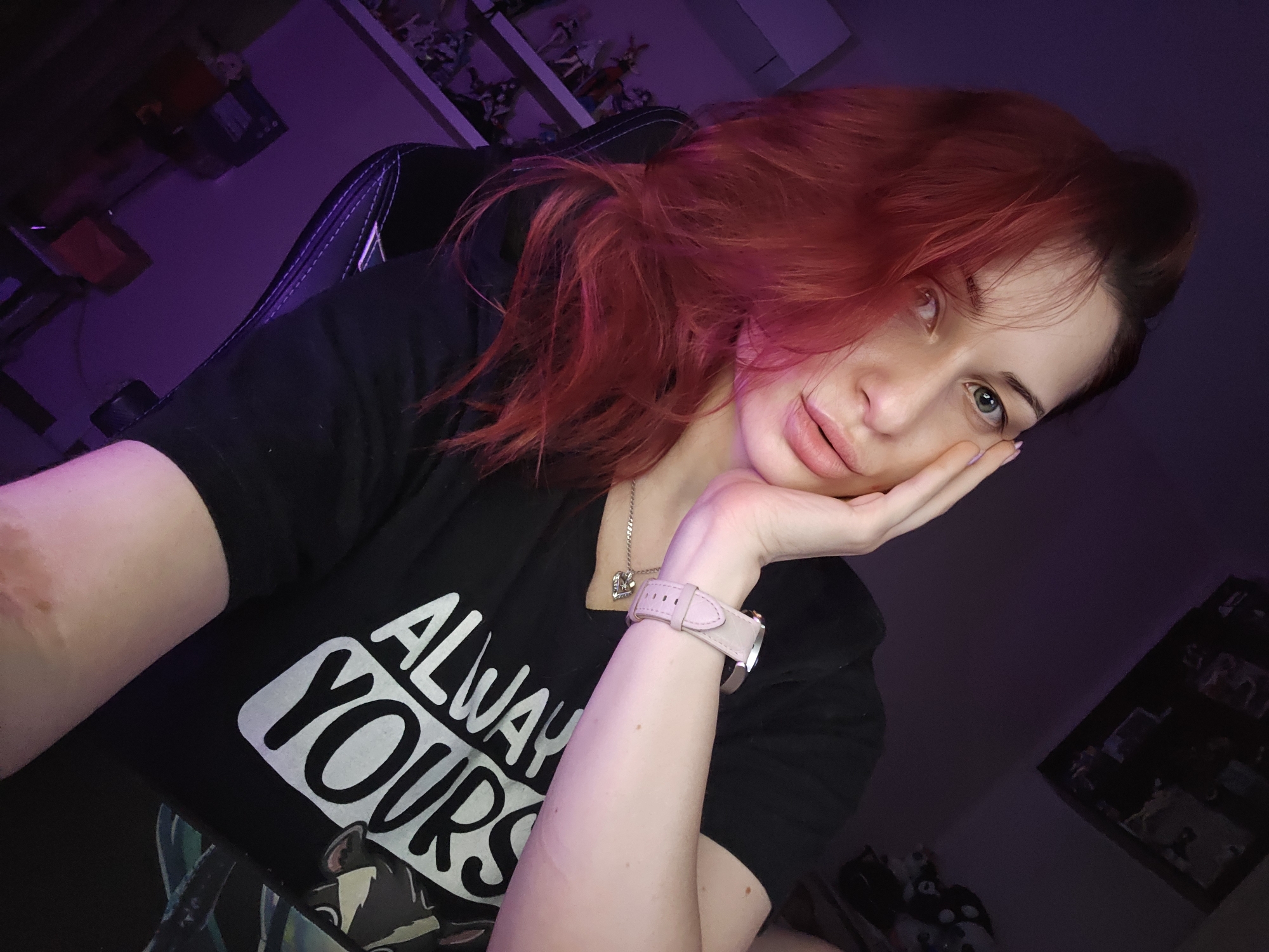 A female individual is depicted in an indoor setting, identified as a personal room in Burgas, Bulgaria. The scene is characterized by dim, purple-hued ambient lighting, emanating from the right side of the frame and illuminating the subject's left profile. The individual has long, reddish-auburn hair, styled with strands framing her face, and light-colored eyes (green/hazel) that directly engage the viewer. She exhibits full lips. Her attire consists of a black short-sleeved t-shirt bearing the white inscription "ALWAY YOURS" across the chest. A silver-toned necklace with a heart-shaped pendant is worn. On her left wrist, she wears a light pink smart or digital watch with a leather-style strap. Her right hand is positioned against her left cheek and jawline, with fingernails painted a pale hue. A noticeable healed scar is present on her right forearm. The individual is seated in a black chair featuring blue accent stitching. The background to the left displays a white shelving unit containing numerous small collectible figurines and a wall-mounted air conditioning unit. The upper right background contains dark, indistinct framed items on the wall. The overall visual tone is moody and artificially lit.